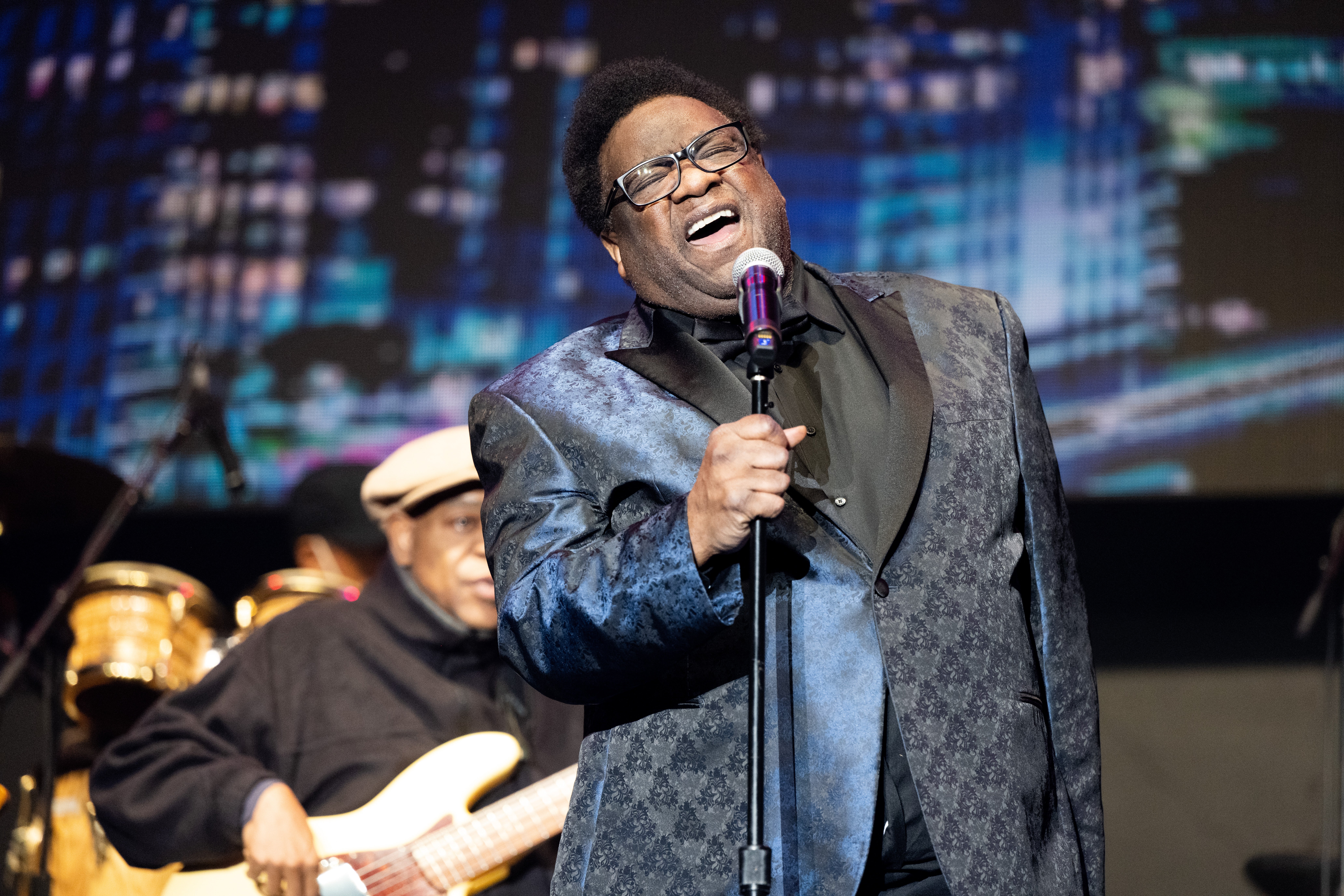 A singer performs passionately on stage, wearing a patterned suit jacket and glasses, with musicians playing instruments in the background