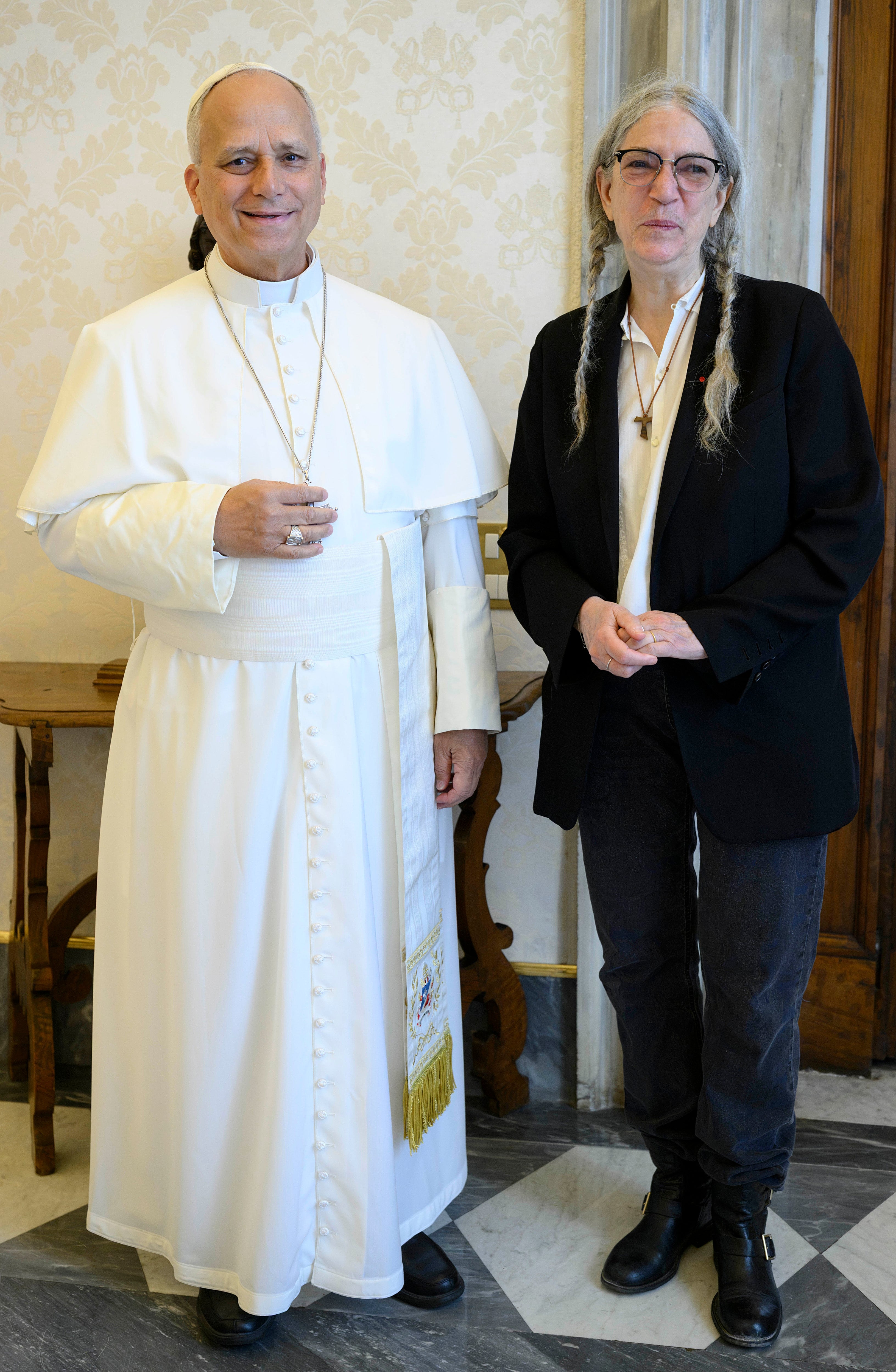 Two individuals stand together; one in traditional religious attire, the other in a suit with long hair. They appear in a formal setting