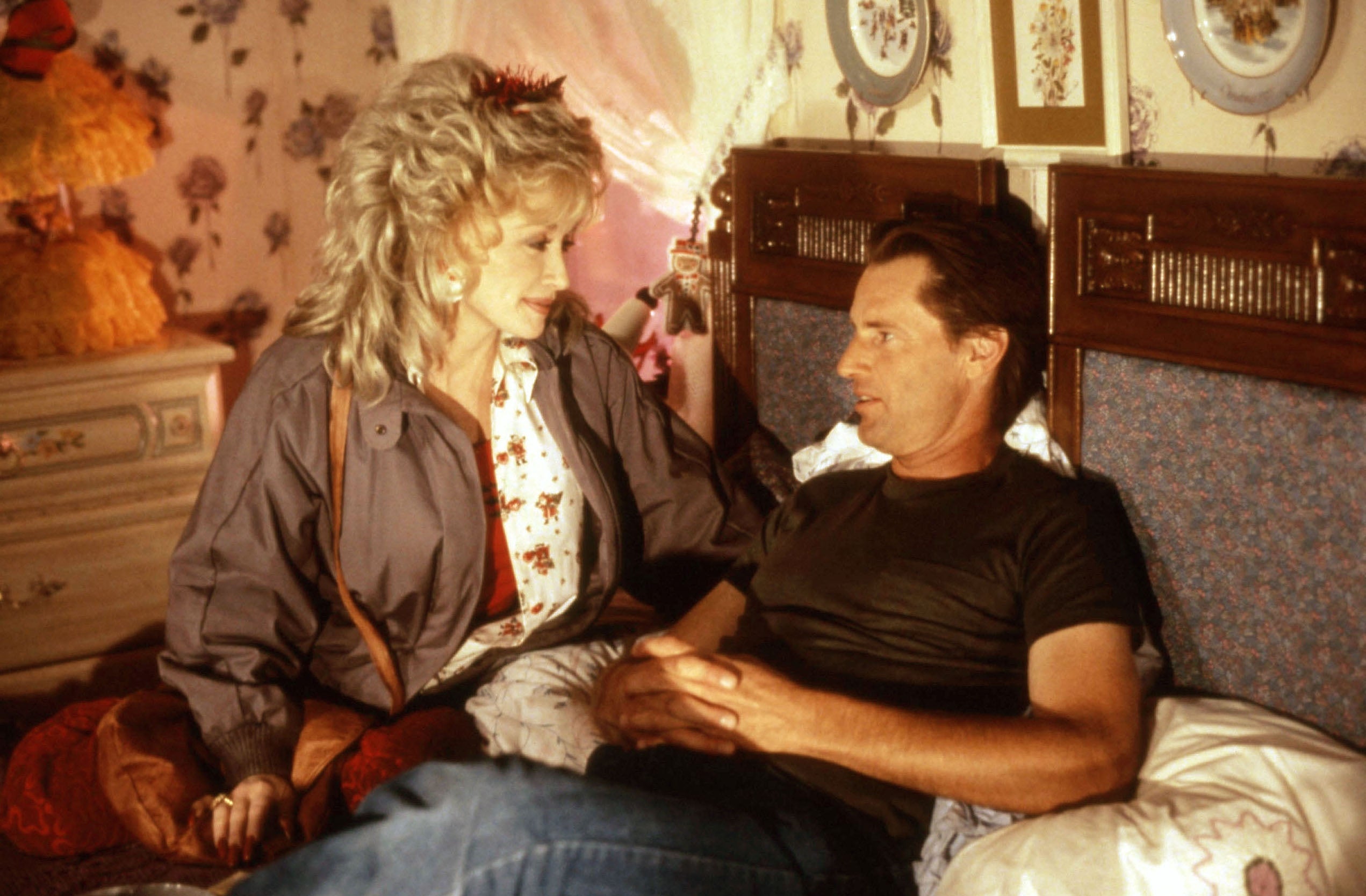 Two people sit on a bed; one wears a casual jacket and blouse, the other a t-shirt. They appear engaged in a conversation