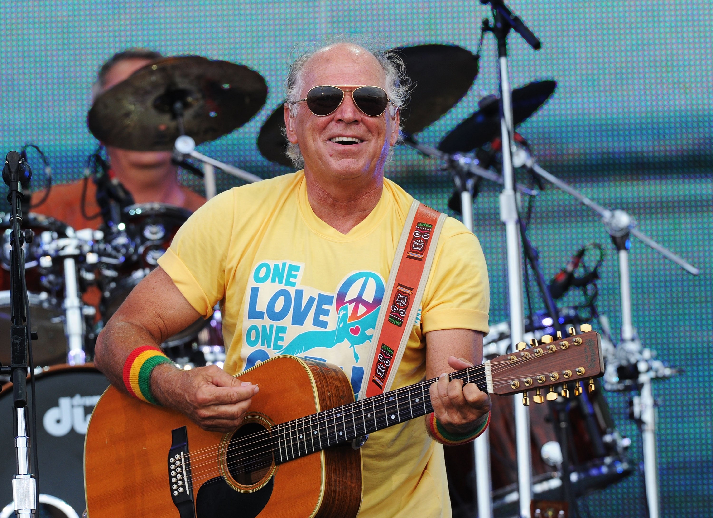 A musician on stage, wearing sunglasses and a yellow graphic t-shirt, plays a guitar with a band in the background