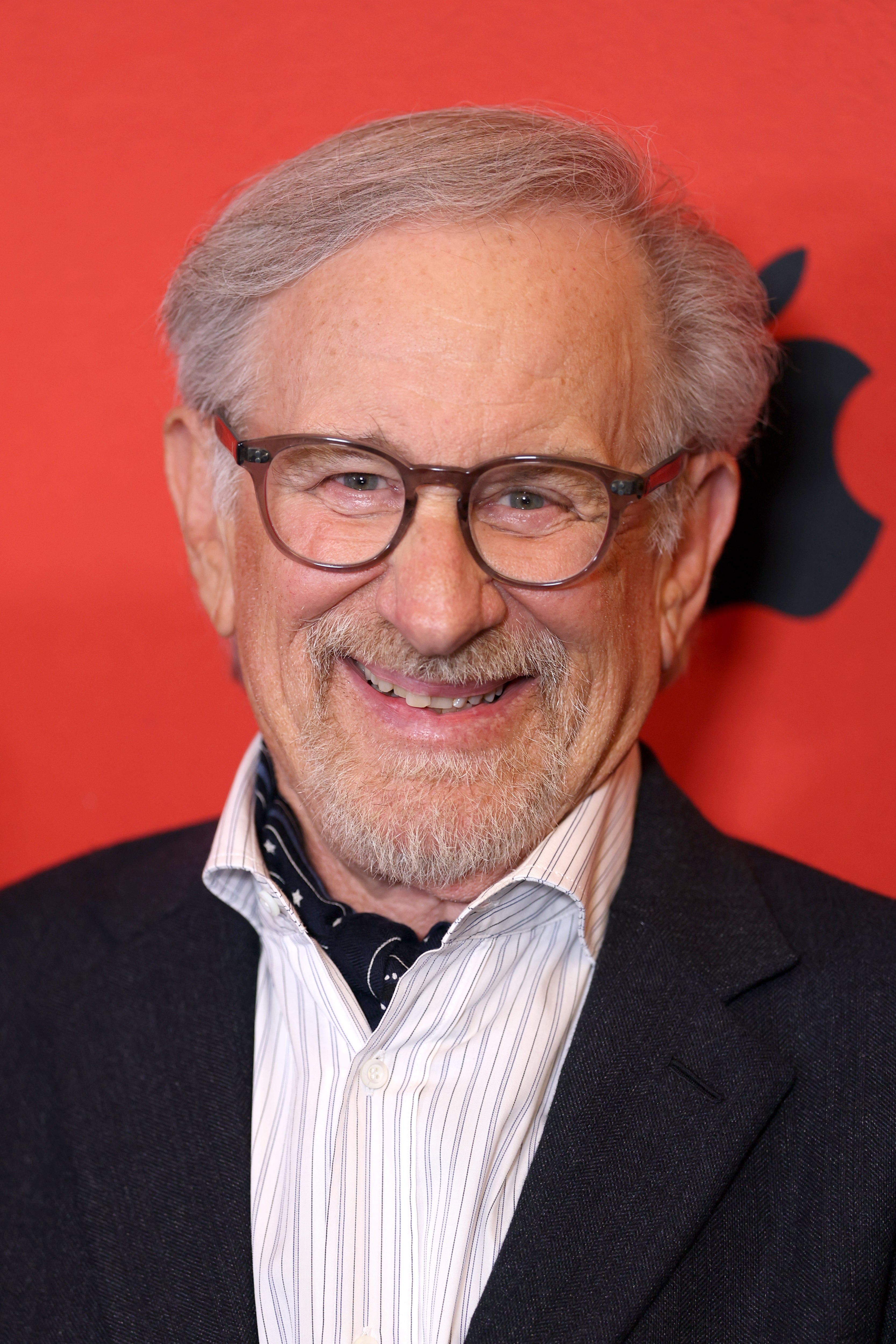 Elderly man in glasses and a suit with a patterned scarf smiles in front of an Apple logo on a red backdrop
