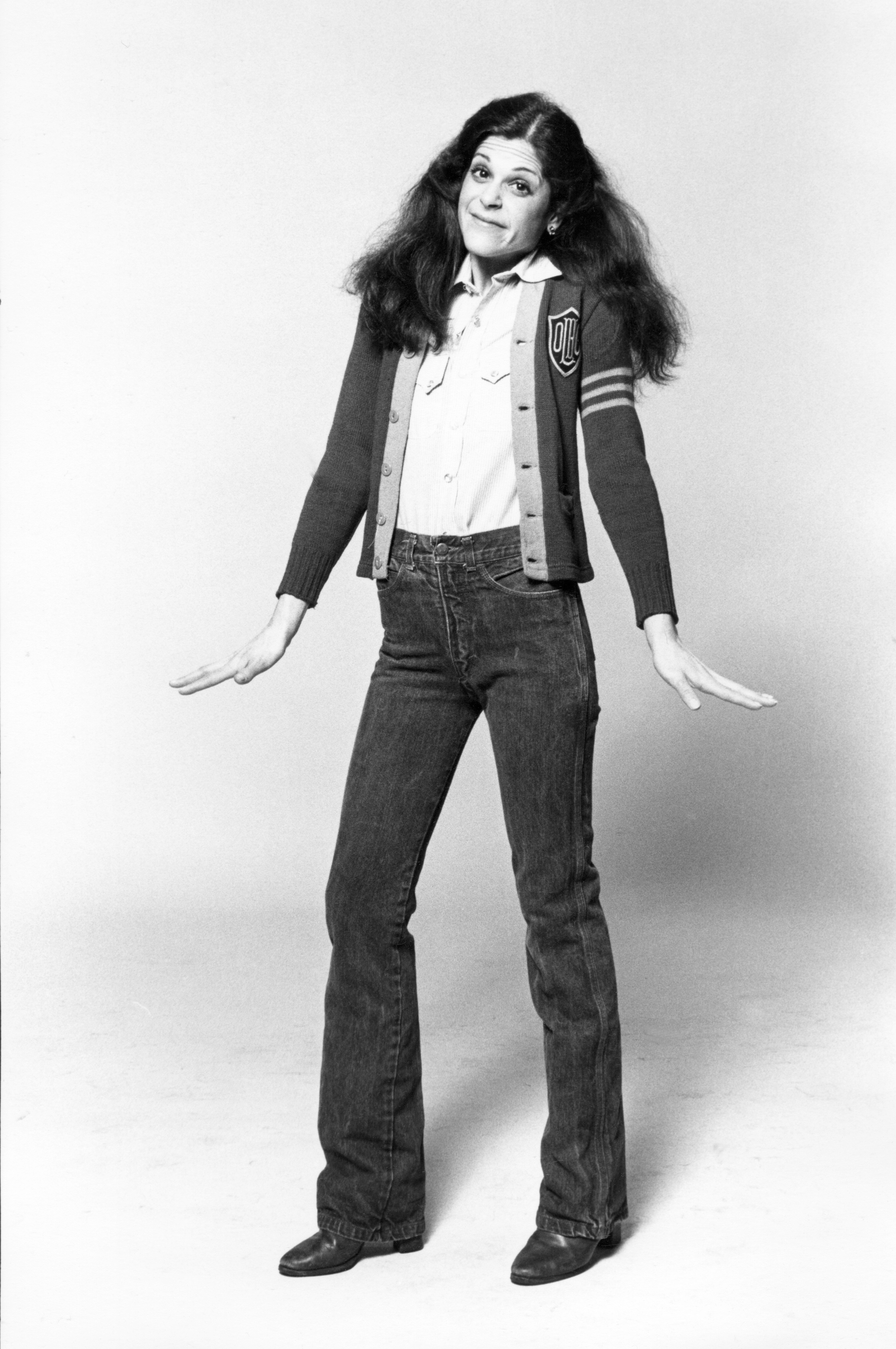 Person with long hair poses playfully, wearing a cardigan, collared shirt, and jeans. Hands are open to the sides, expressing a casual demeanor