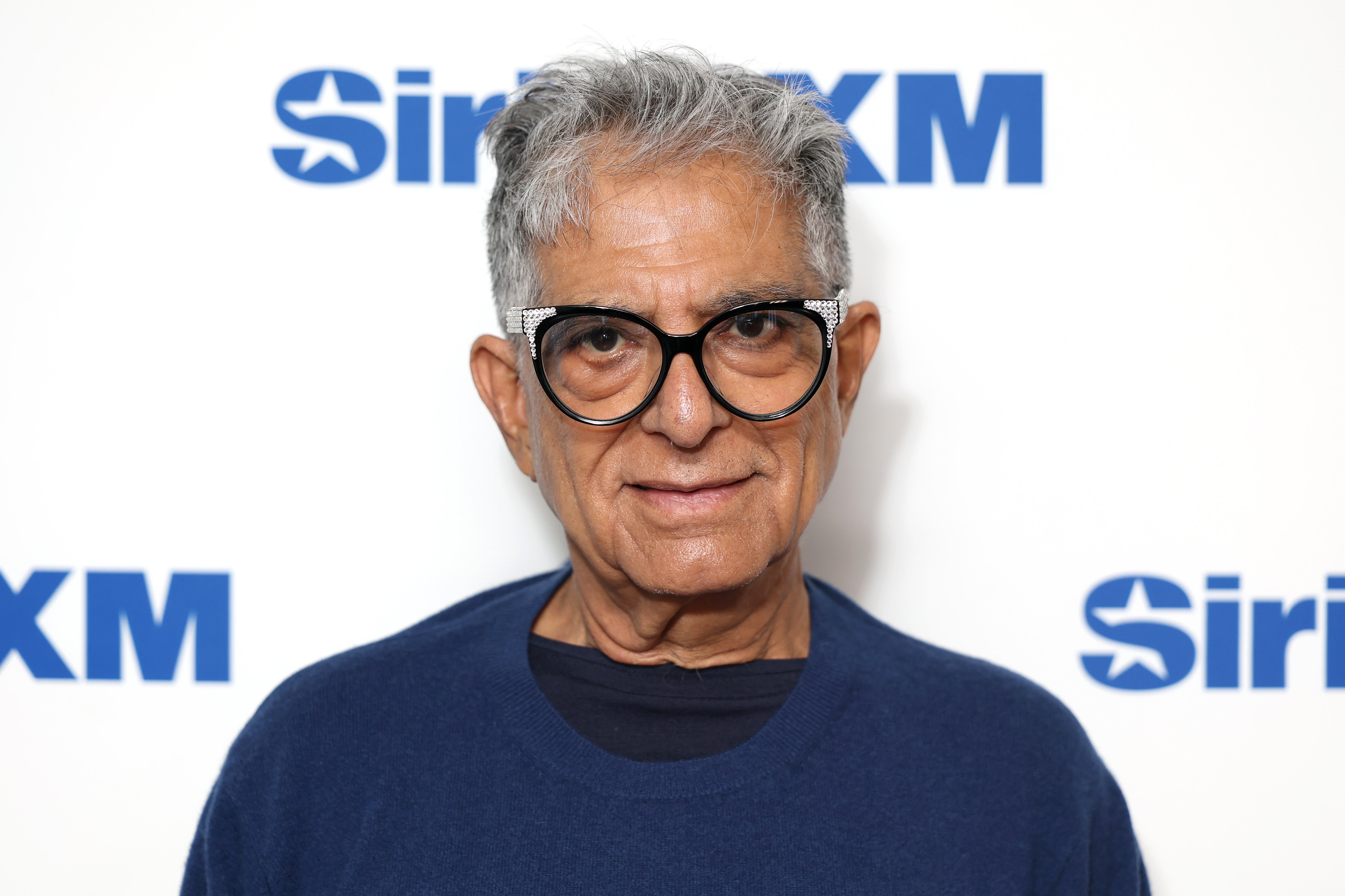A person is posing in front of a wall with the SiriusXM logo, wearing glasses and a simple top