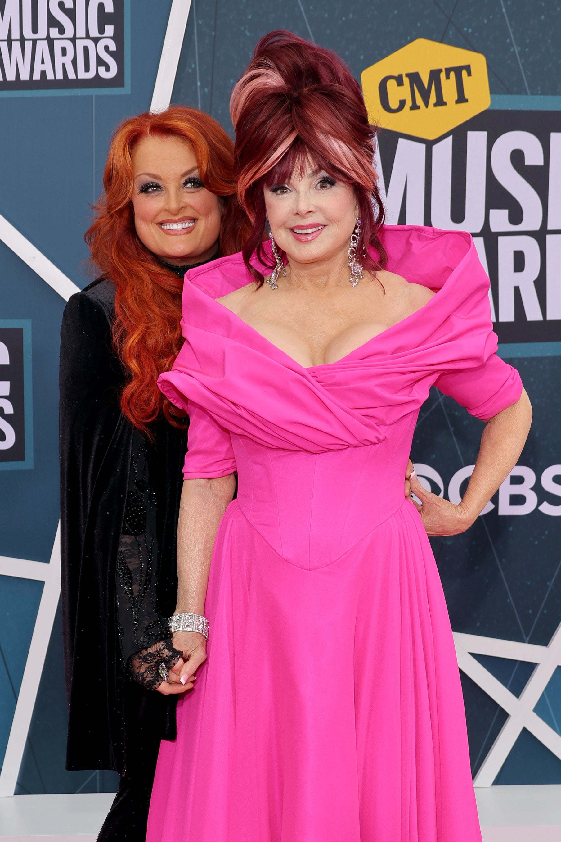 Two women stand on the CMT Music Awards red carpet. One wears a pink dress with a wide collar; the other has long hair and wears a black outfit