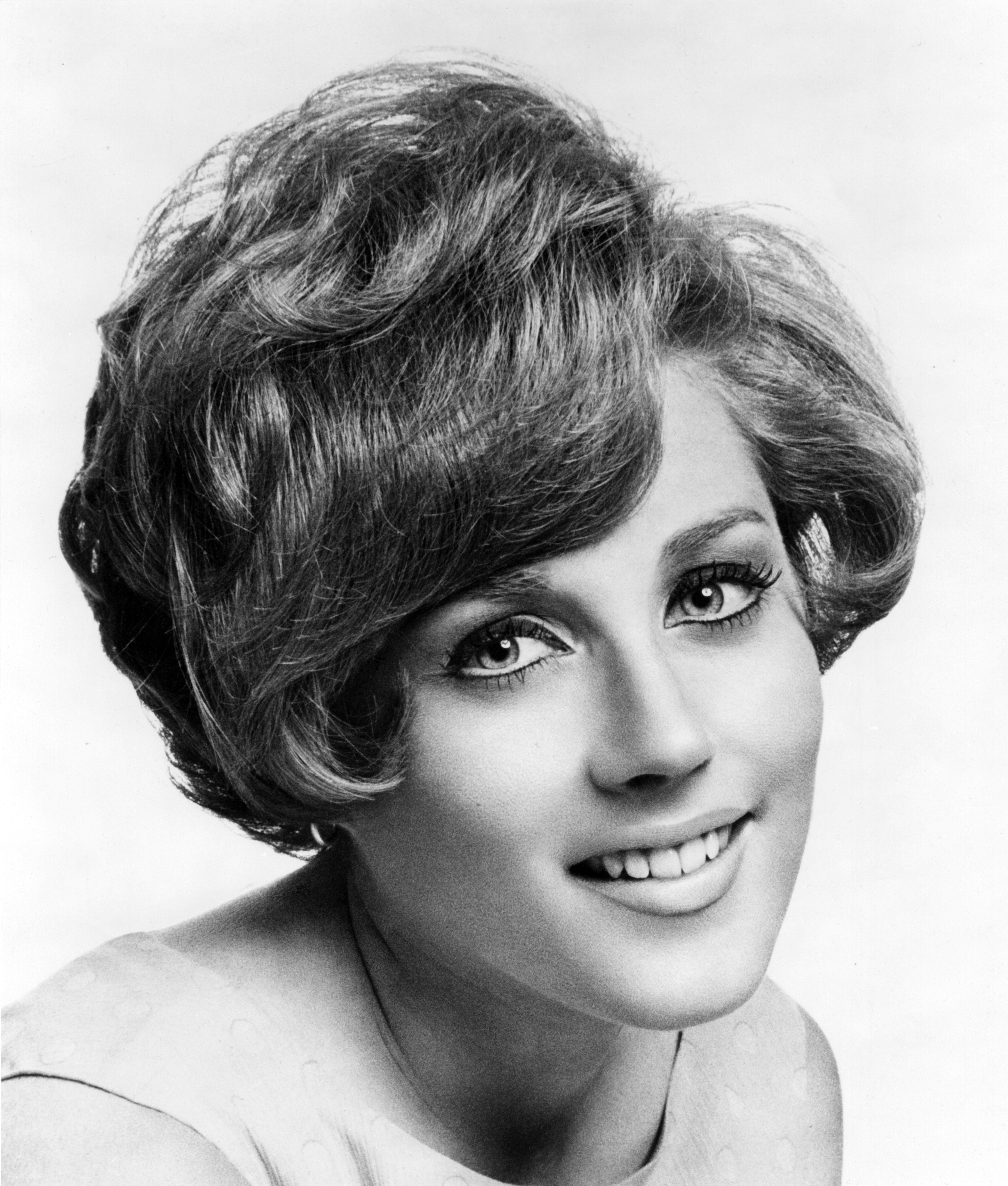 Person with a vintage hairstyle and a smile, wearing a simple top, in a classic black-and-white portrait