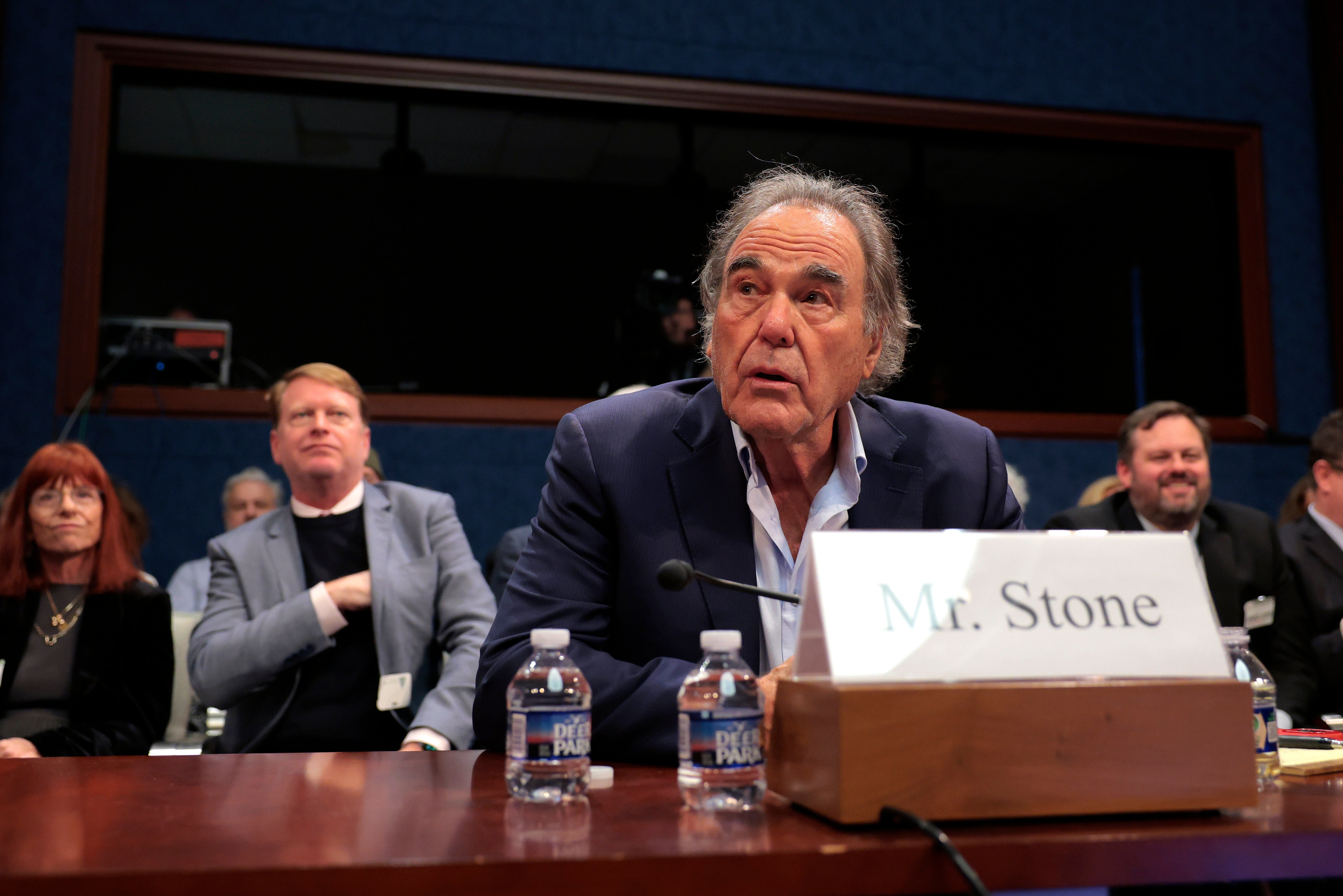 A man in a suit sits at a hearing table, labeled as "Mr. Stone," with people seated behind him in a formal setting