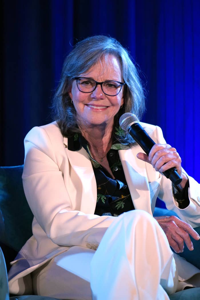 A person in a white suit is seated, holding a microphone. The individual has glasses and appears to be speaking at an event