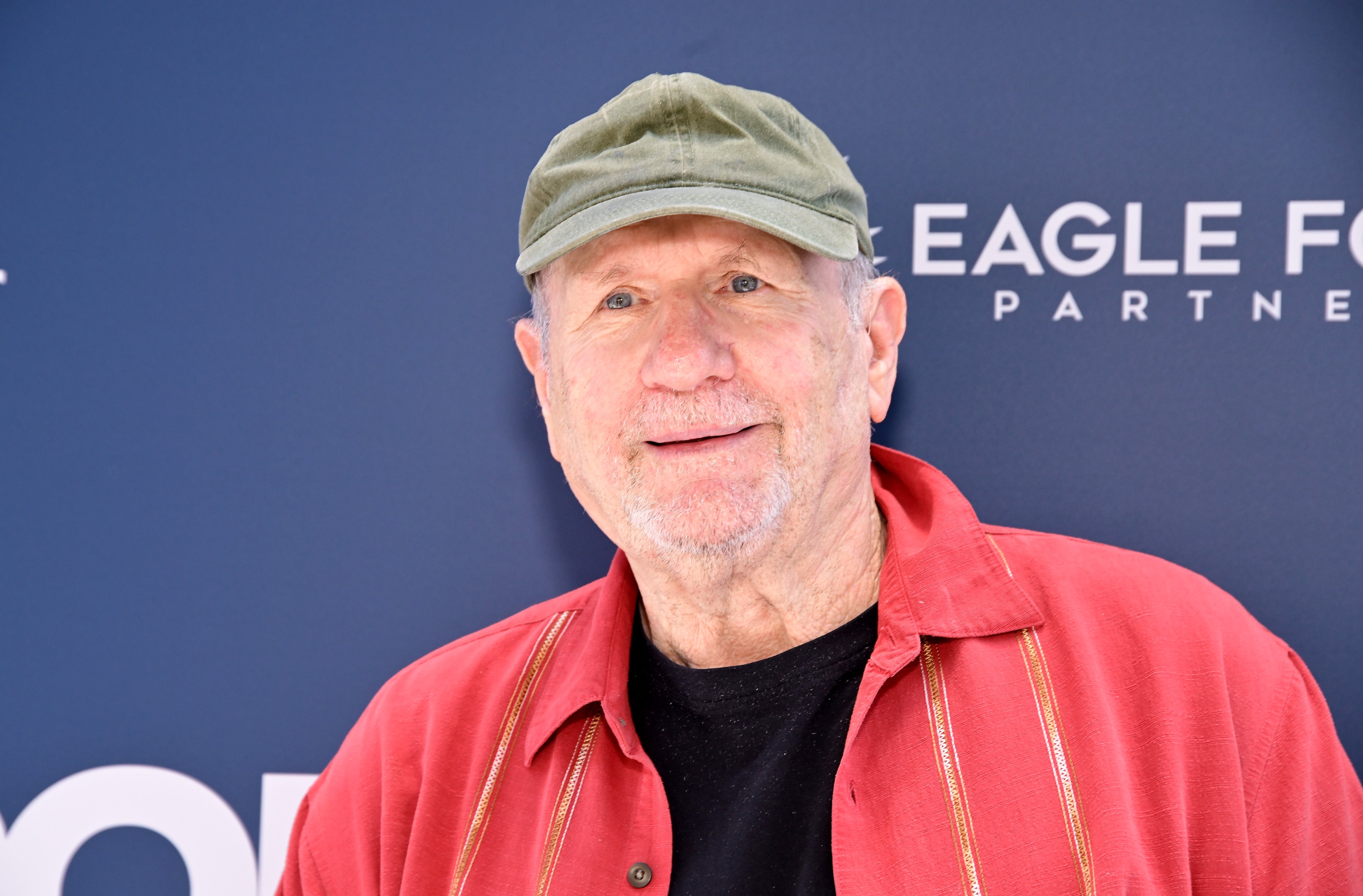 A person in casual clothing, wearing a hat and red jacket, smiles at a celebrity event backdrop