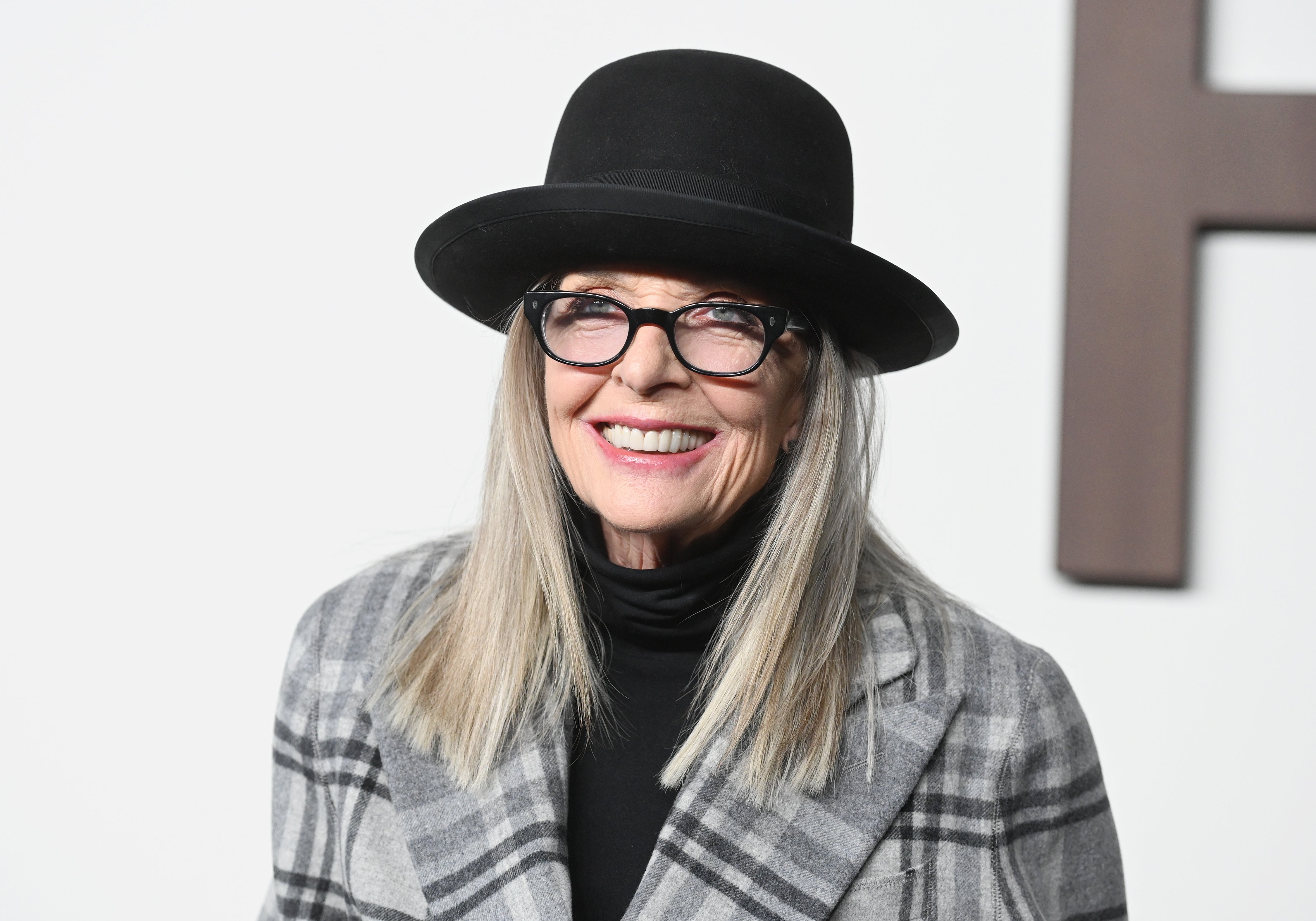 Person wearing a plaid coat, black turtleneck, and wide-brim hat, smiles on the red carpet with glasses