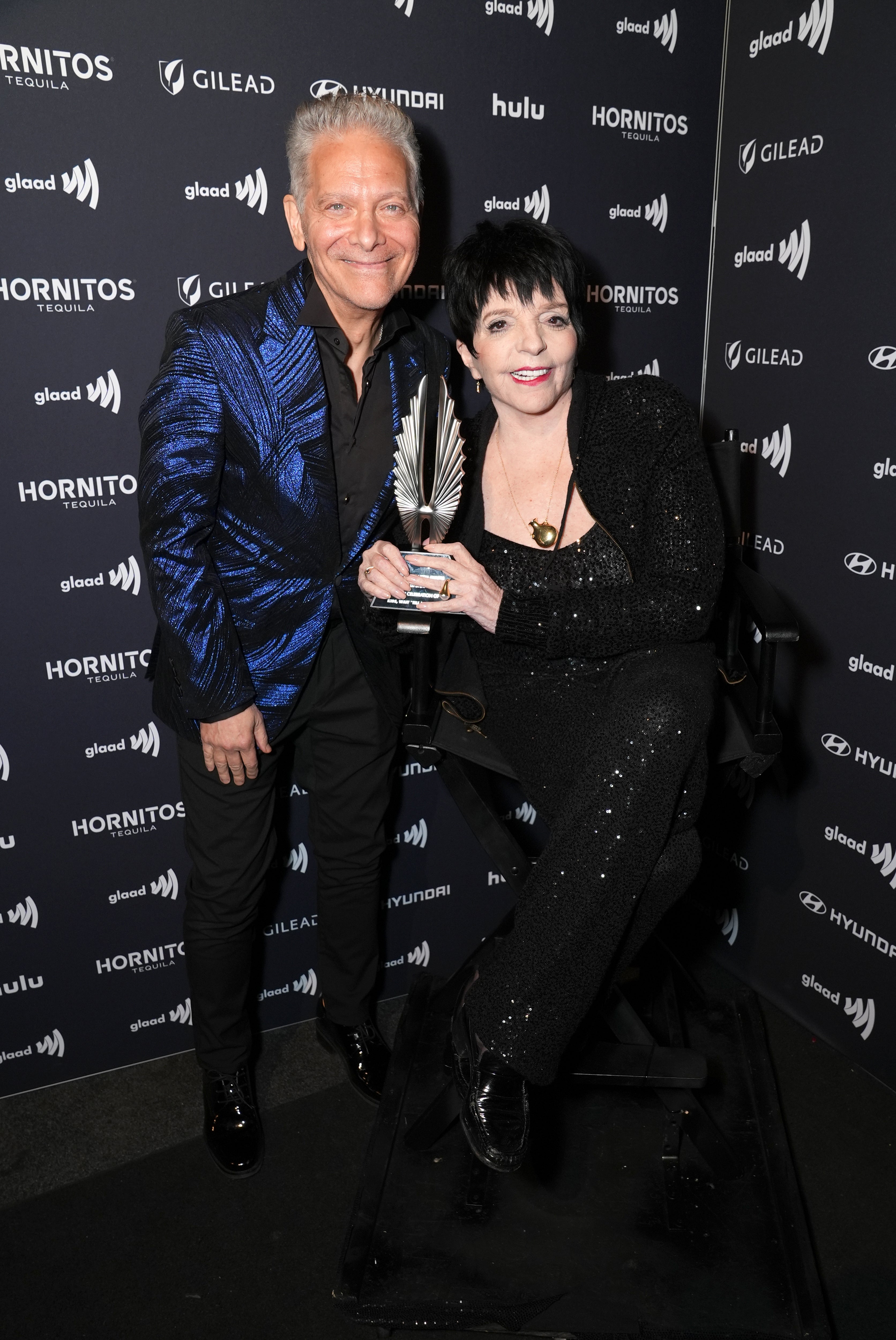 Two people pose at an event, one holding an award. They're dressed in elegant, glittering outfits suitable for a formal occasion
