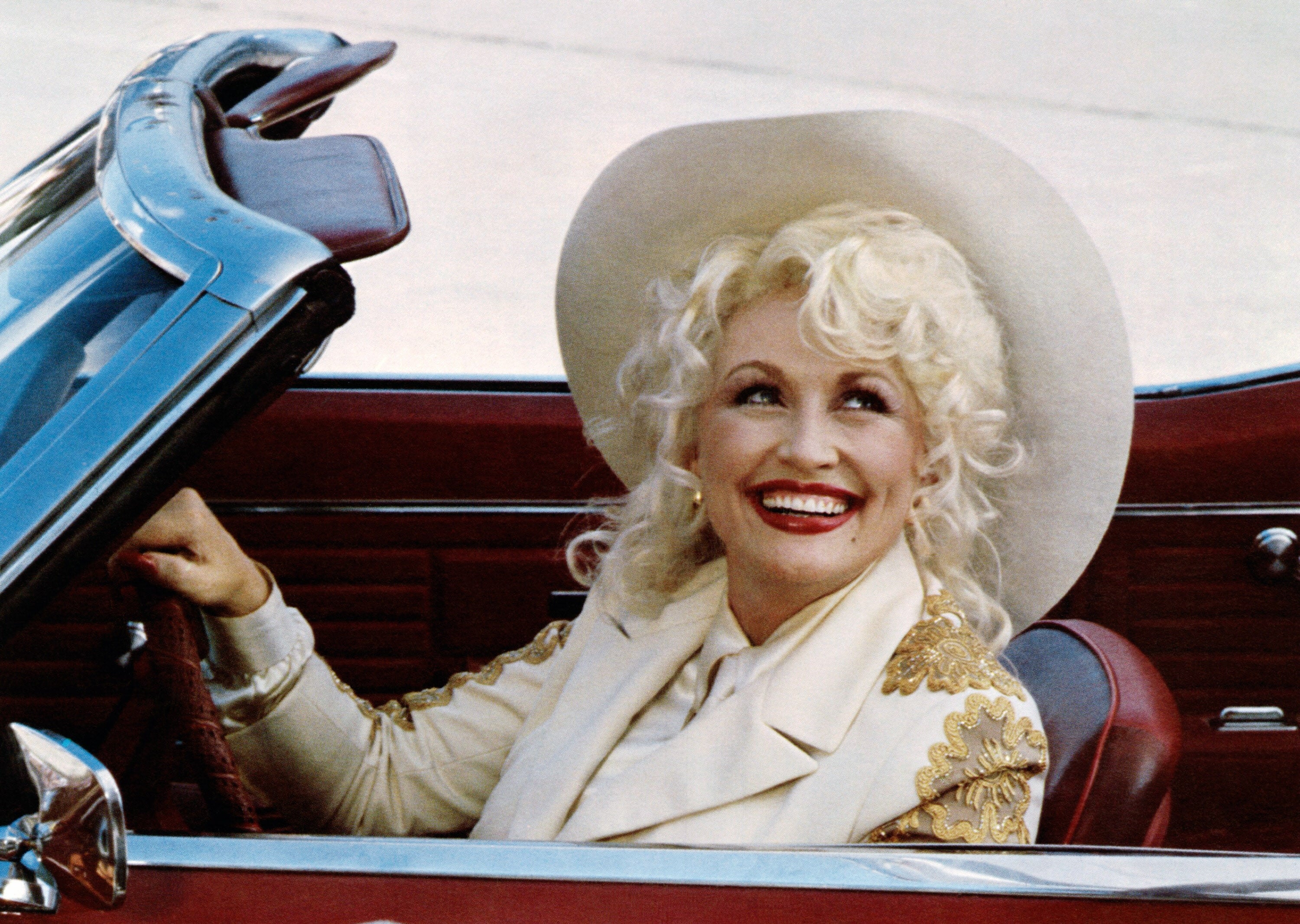 Person smiling in a convertible, wearing a wide-brimmed hat and an embellished jacket, exuding a warm, elegant style