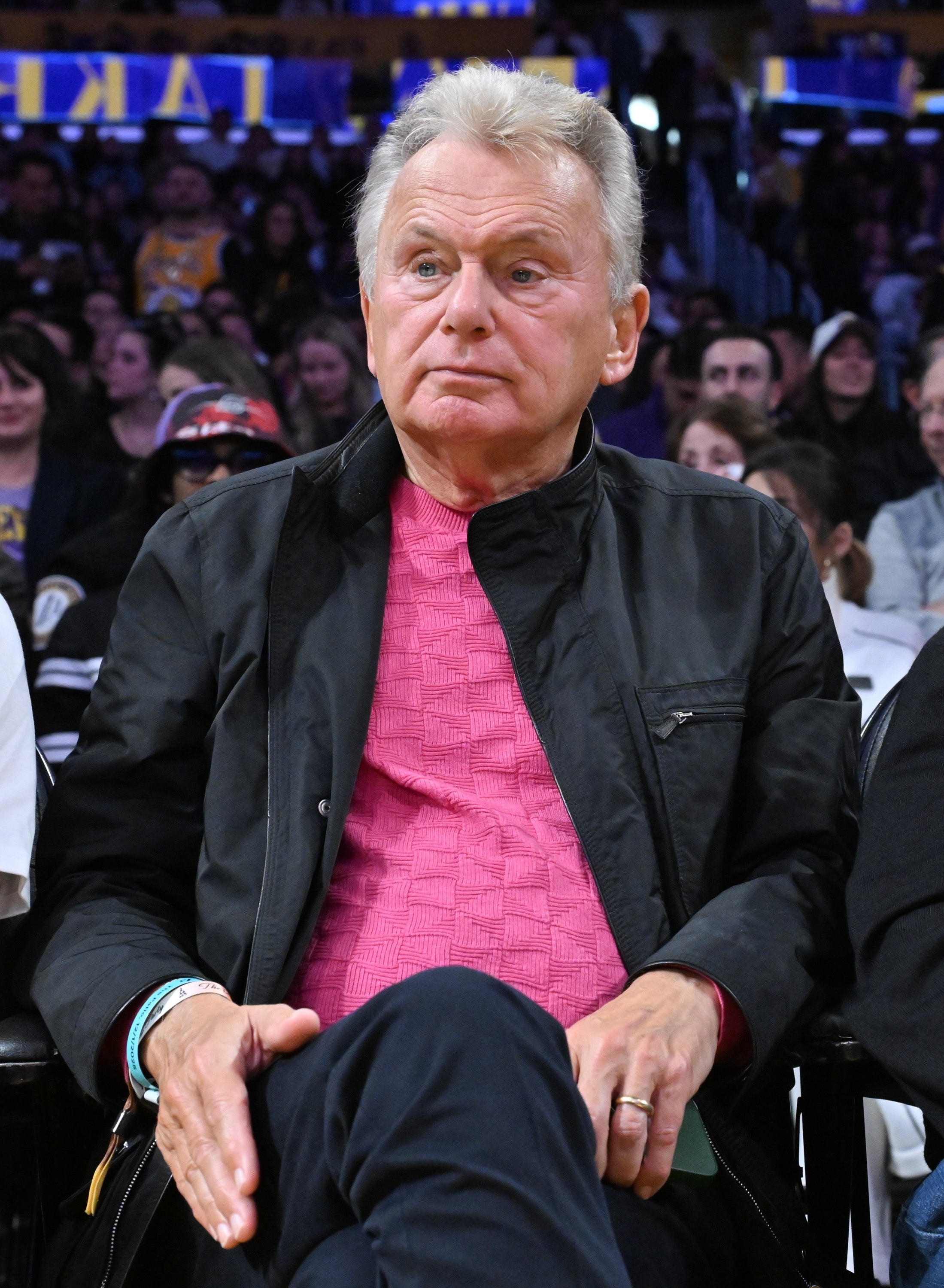 A man in a casual pink sweater and black jacket sits at a public event, surrounded by other attendees