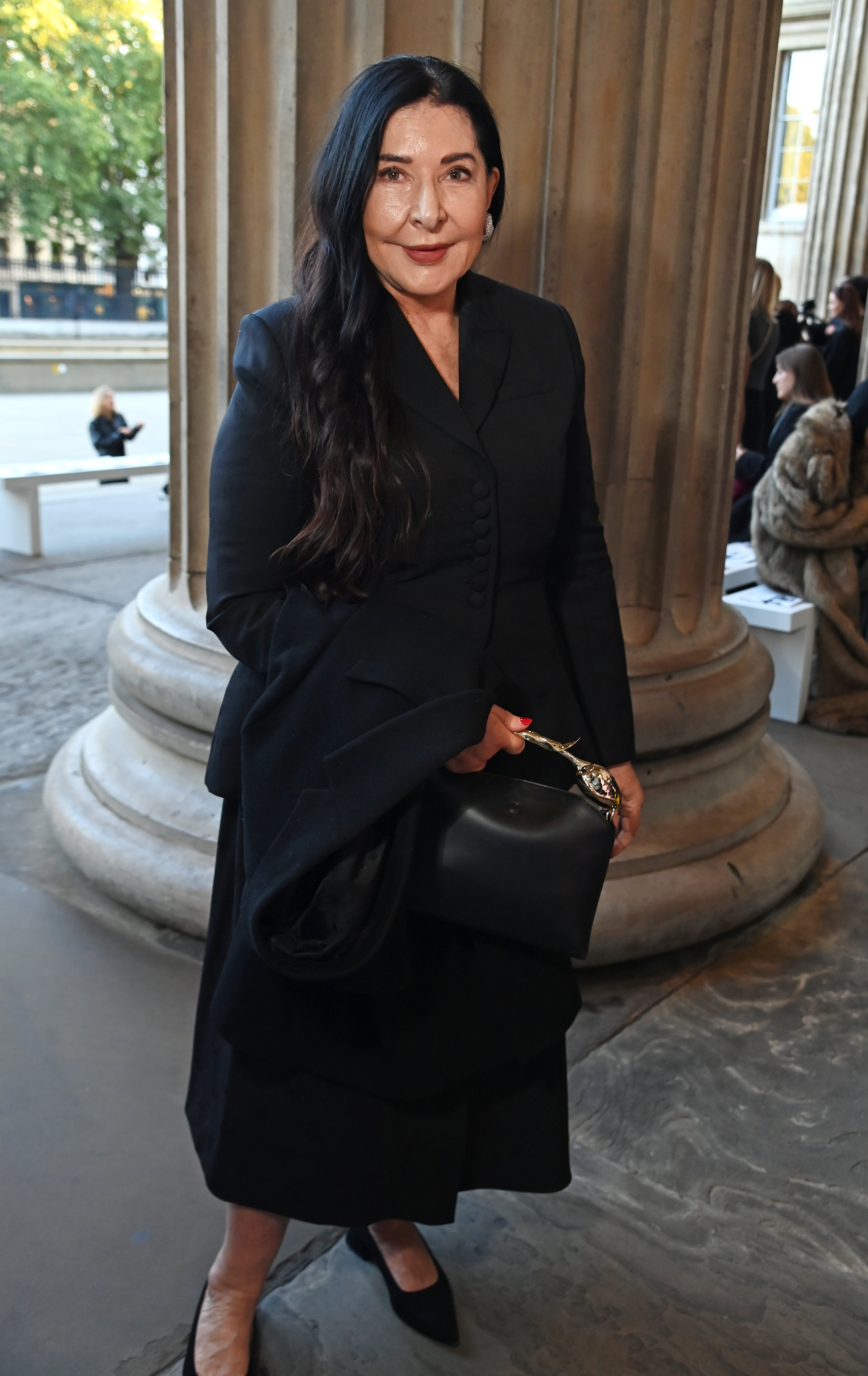 Person in a sleek black coat and matching dress, carrying a black bag, standing near columns at an outdoor event