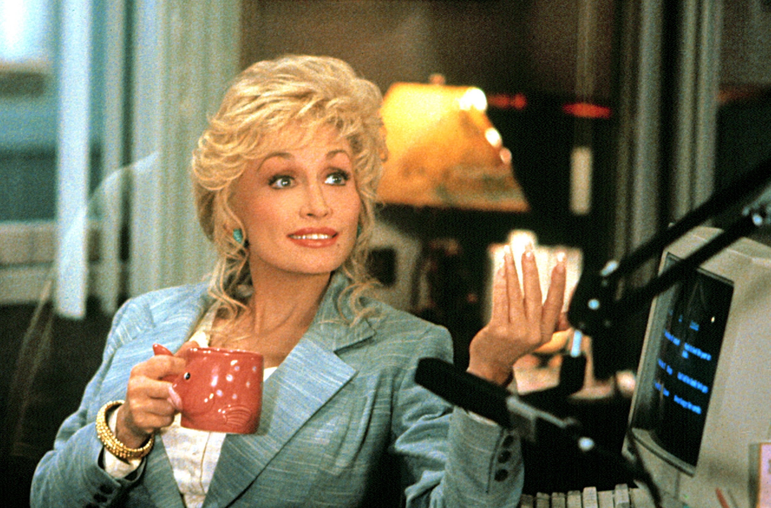 A person in a business suit holds a mug, gesturing with their other hand, seated at a desk with a computer