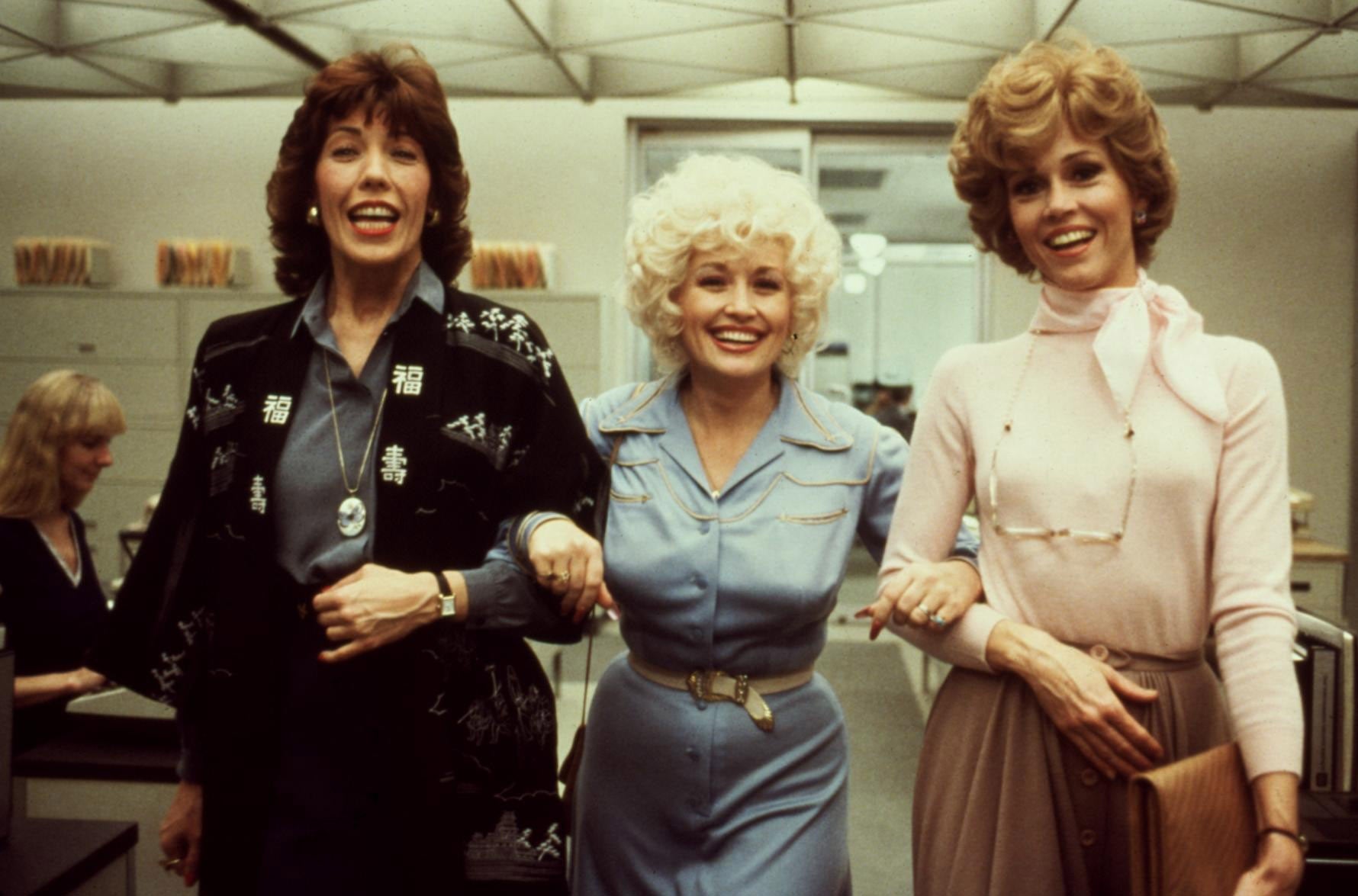 Three women smiling and linking arms in an office setting, wearing business-style attire from the late 20th century