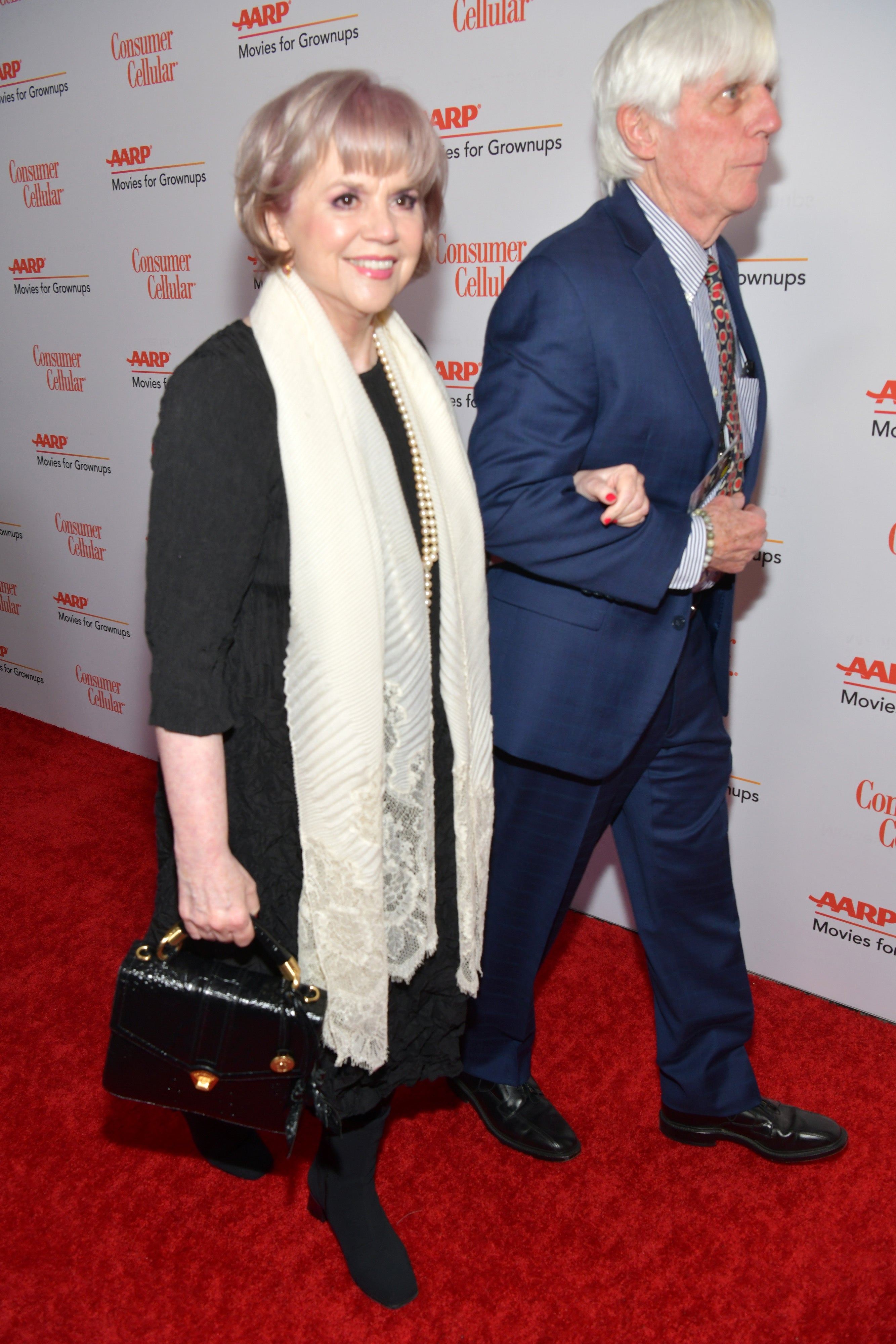 Woman in elegant black dress with lace scarf and man in a blue suit walking on a red carpet event backdrop