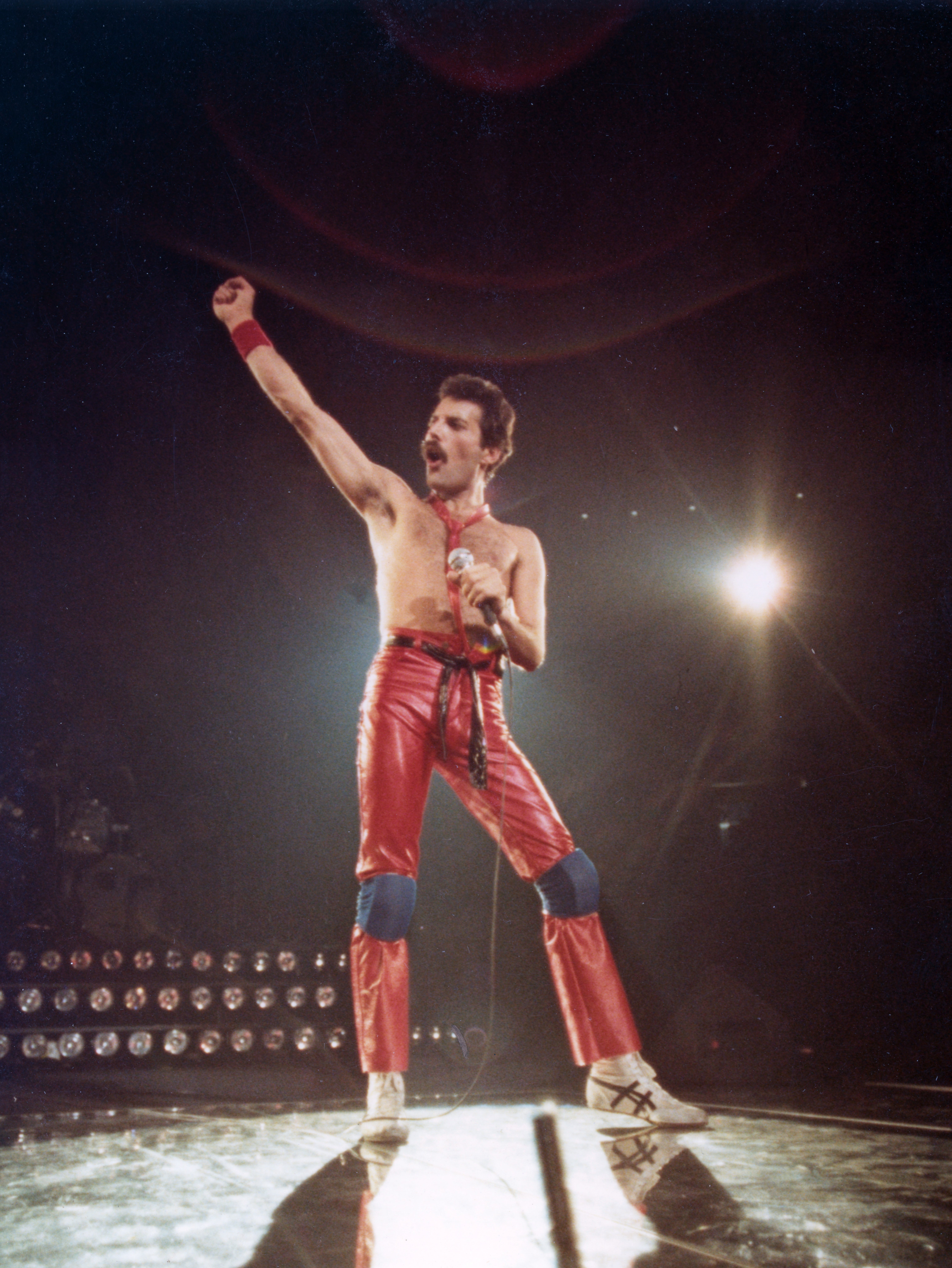 Performer on stage in dynamic pose, wearing shiny red pants and holding a microphone, with stage lights creating a dramatic backdrop