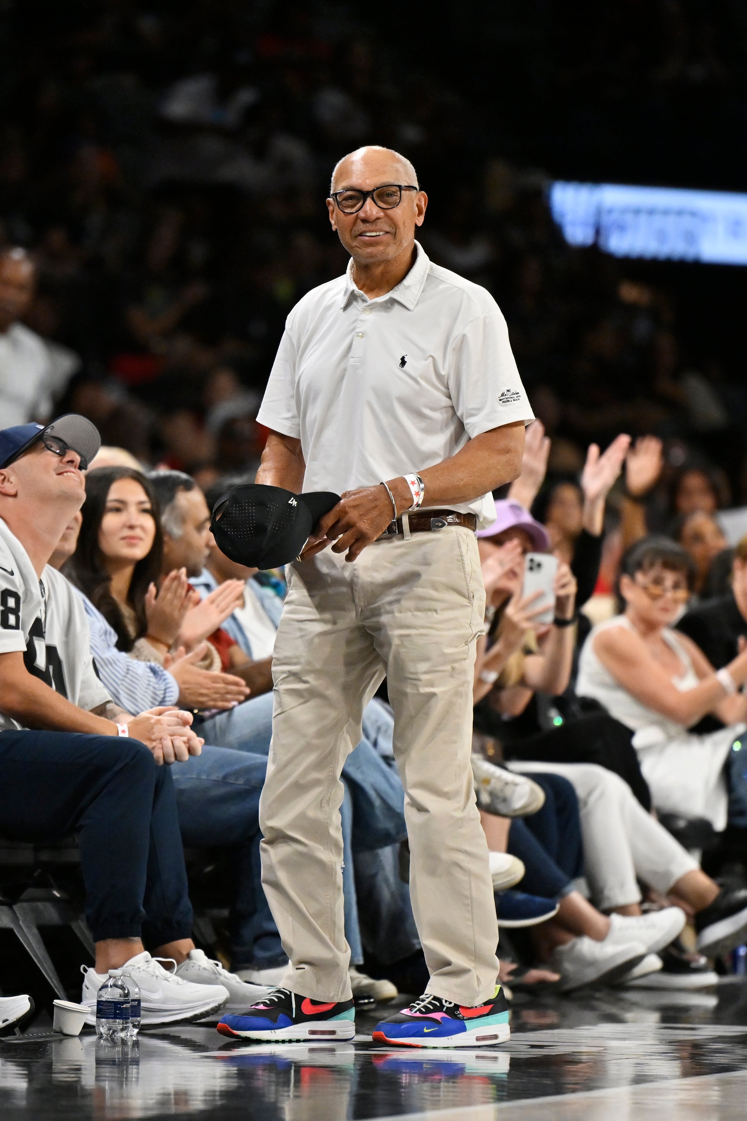 Person wearing casual attire with a polo shirt, holding a cap, standing in a sports arena with audience clapping
