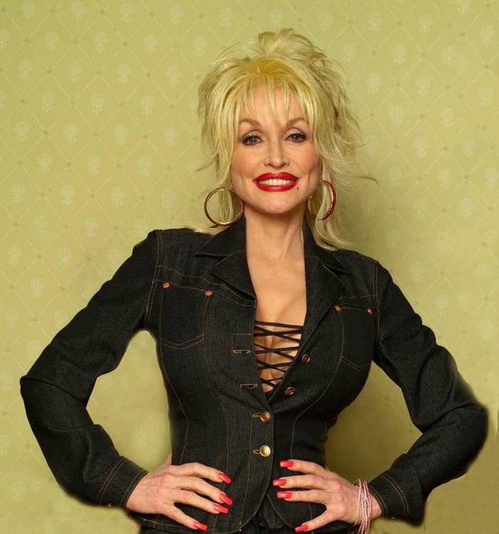A person with styled hair and large earrings is wearing a fitted denim outfit with a lace-up front, smiling with hands on hips