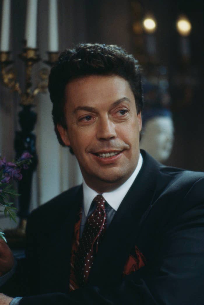 Actor smiling in a formal setting, wearing a classic suit with a polka-dot tie, seated indoors with dim lighting