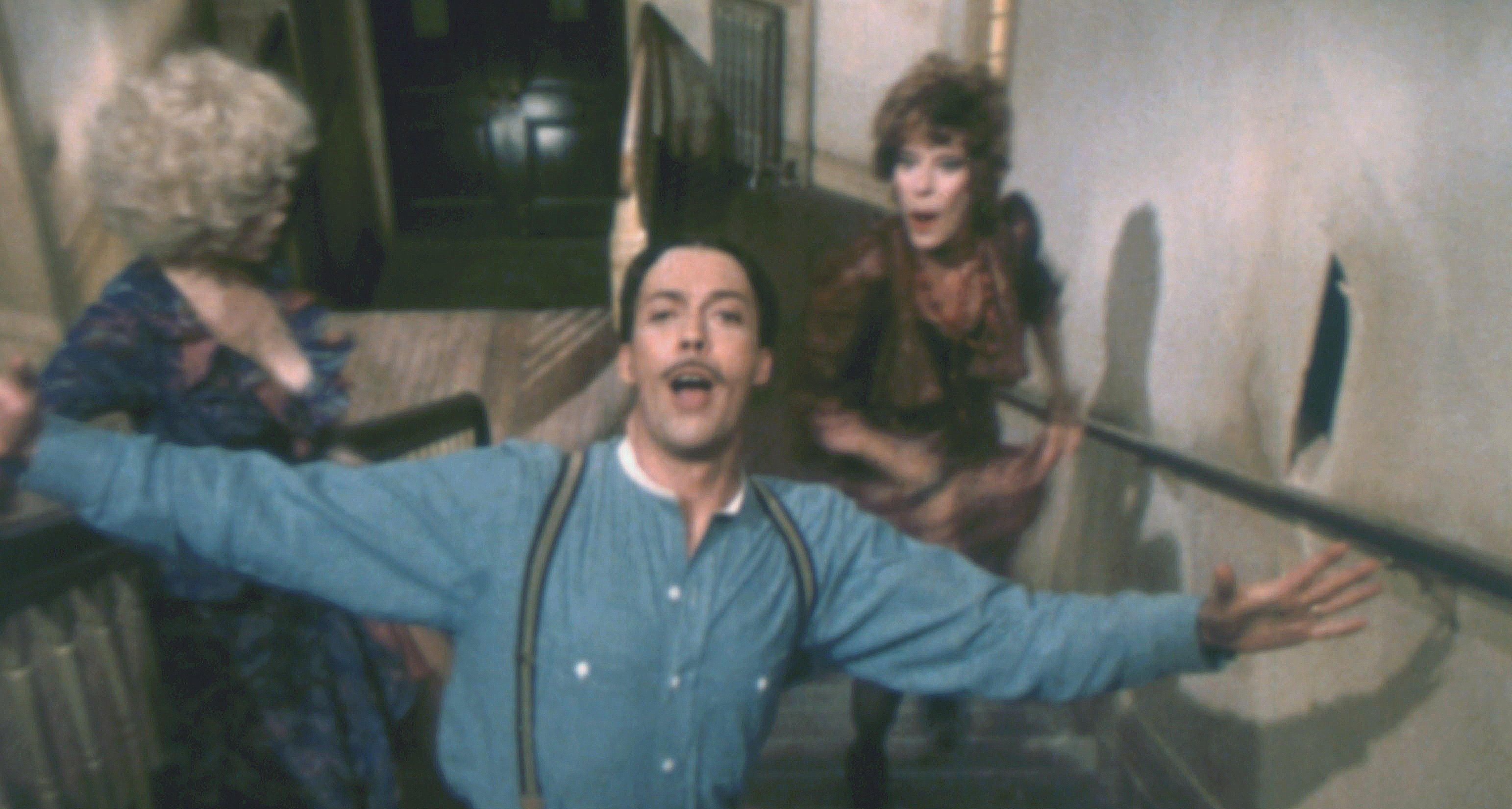 A person with a mustache and suspenders joyfully poses on a staircase, with two people in the background