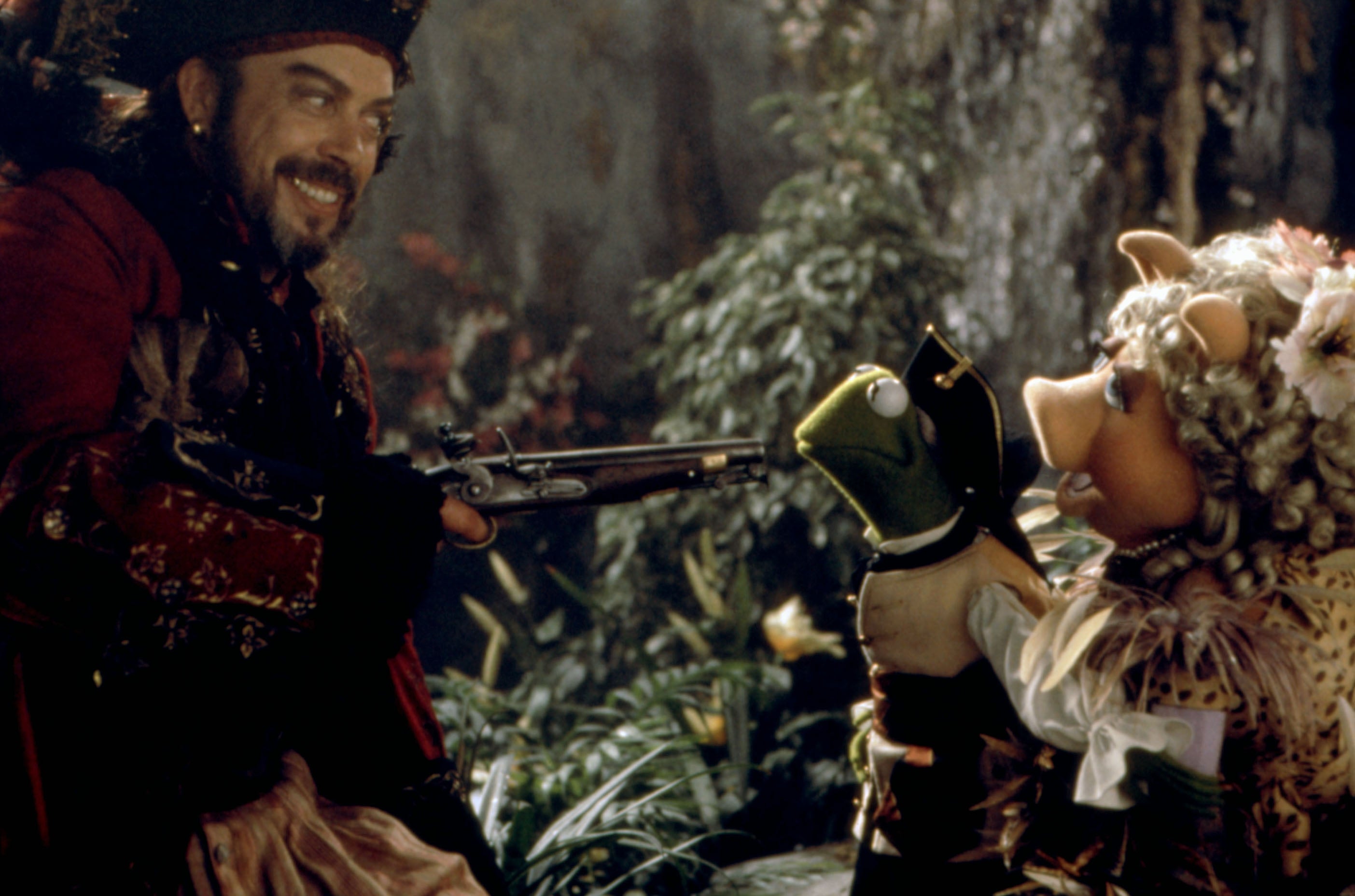 A pirate points a musket at a frog puppet, held by a glamorously dressed pig puppet in a forest scene