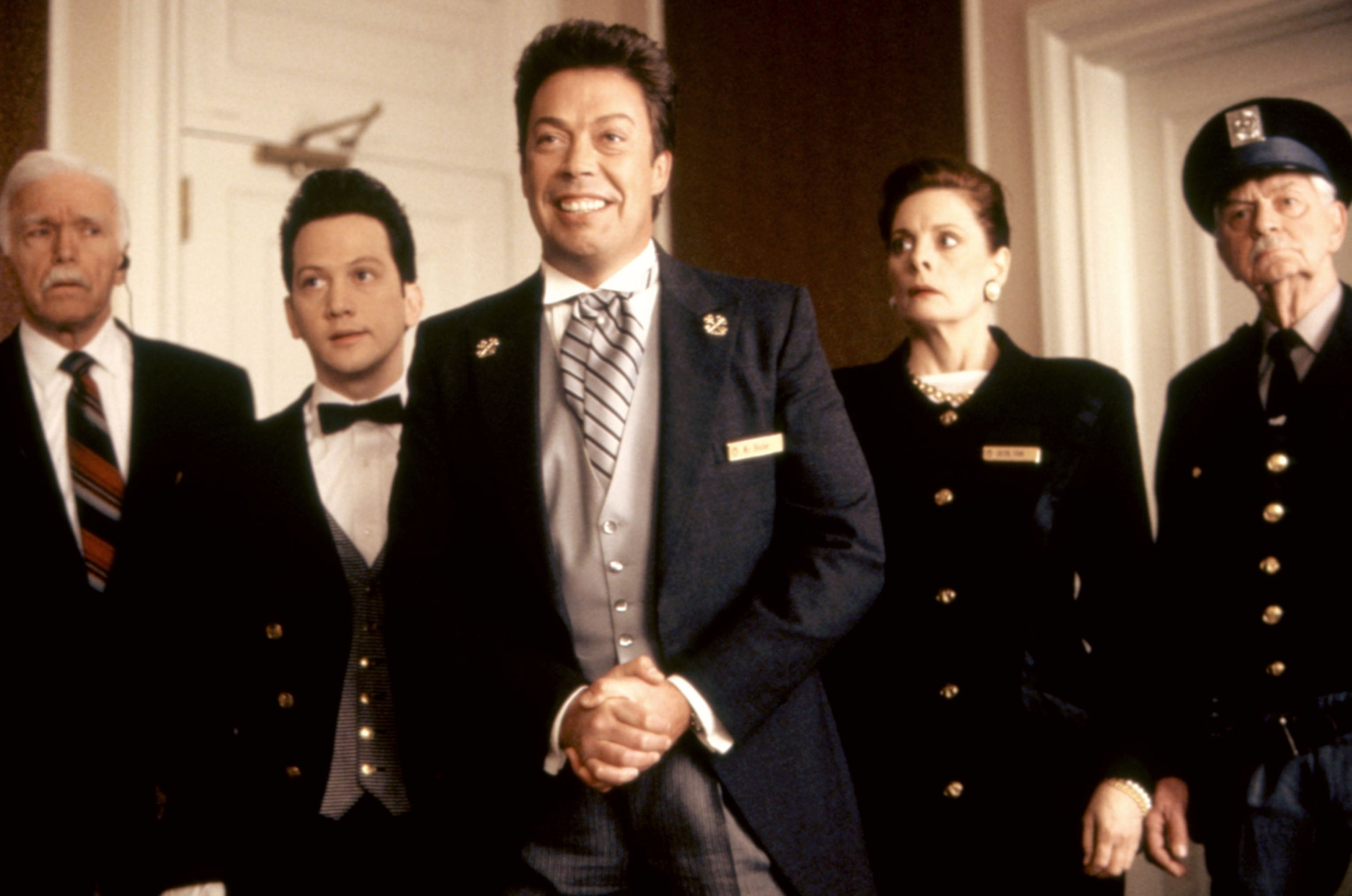 Five people in formal attire, including a security guard, stand indoors, appearing attentive and slightly surprised