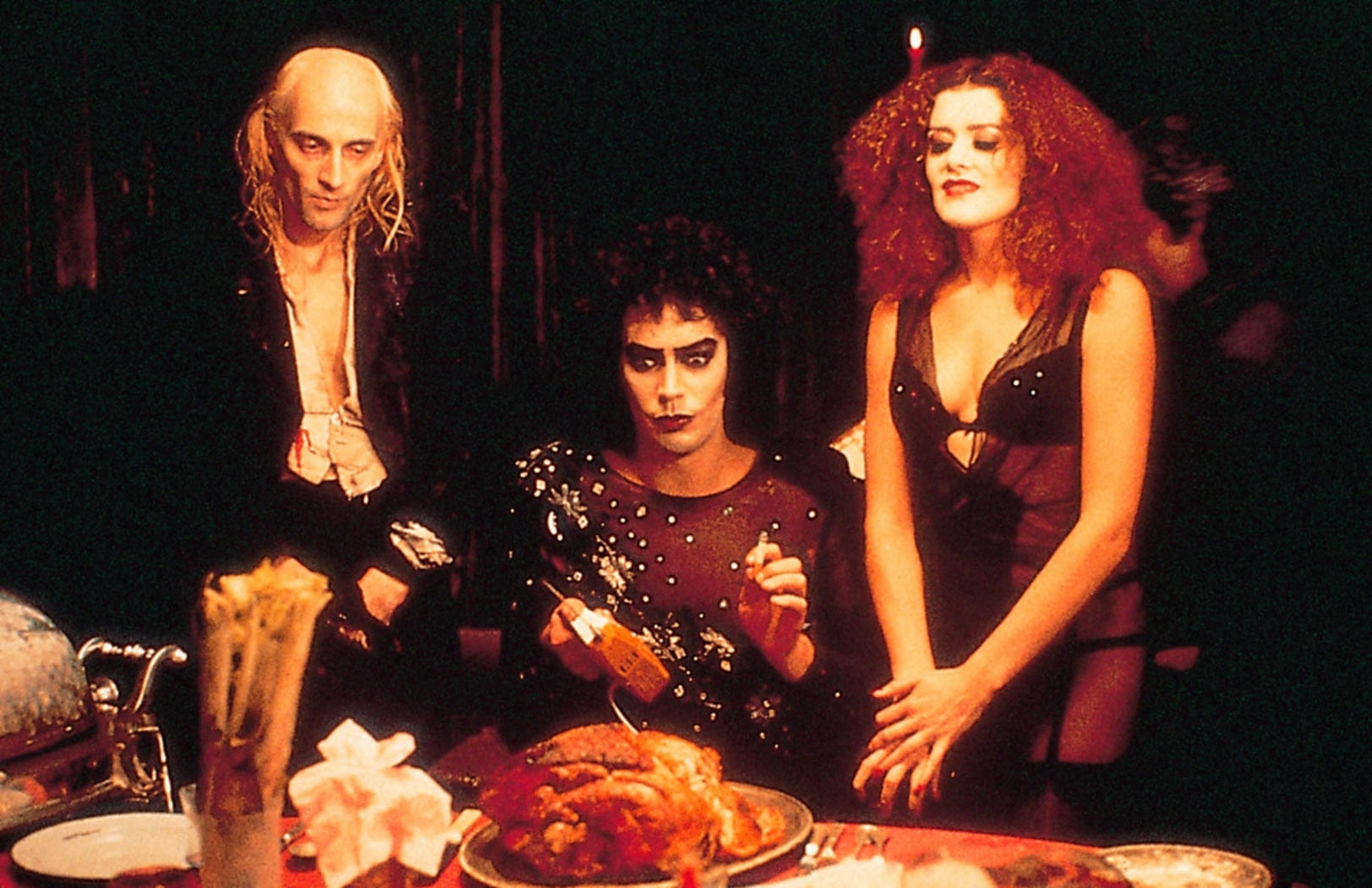Three people in dramatic, gothic-style clothing stand around a dining table with a cooked turkey centerpiece