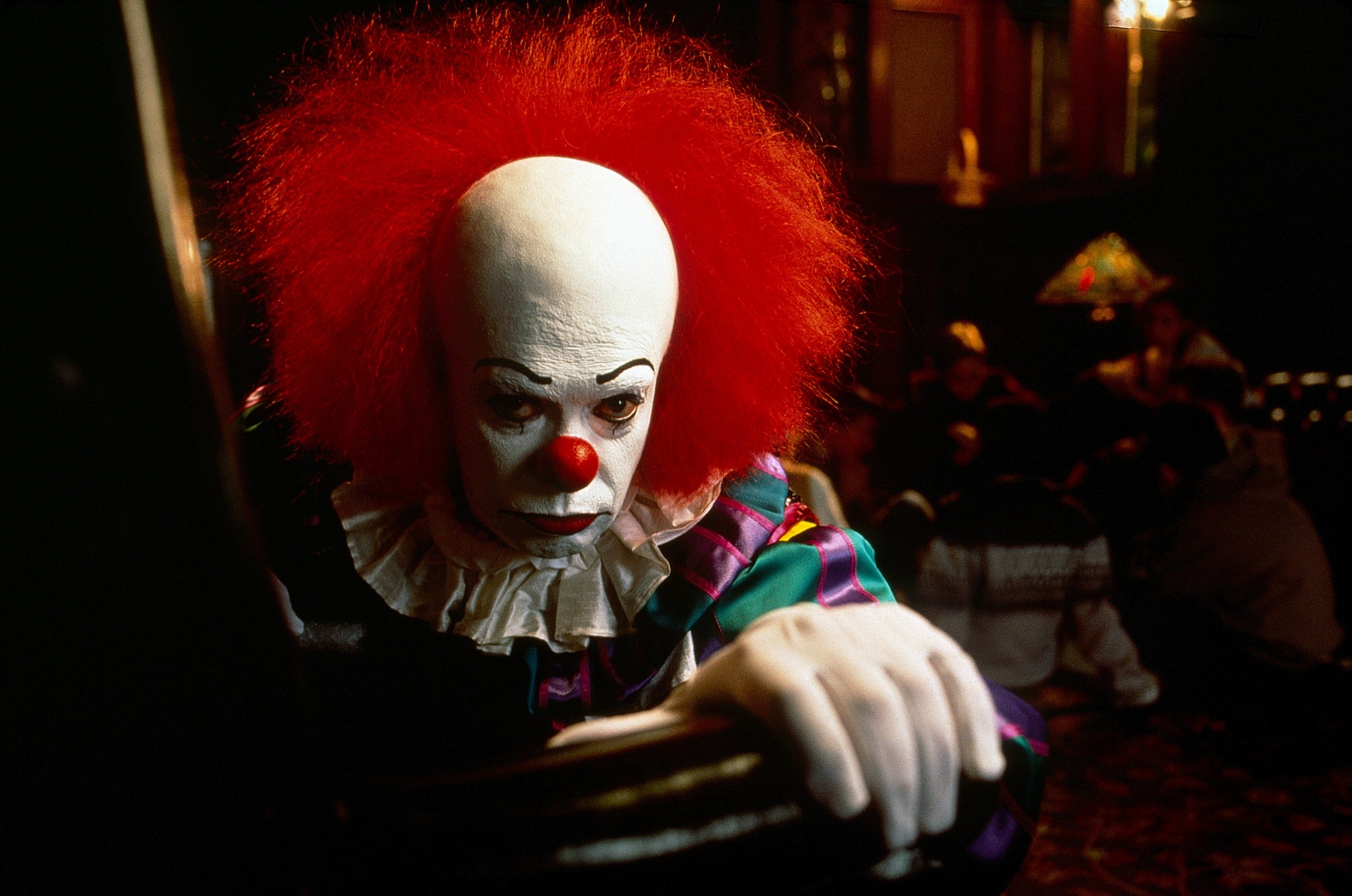 A clown with a red nose and white face paint looks menacingly at the camera, wearing a ruffled collar and colorful costume