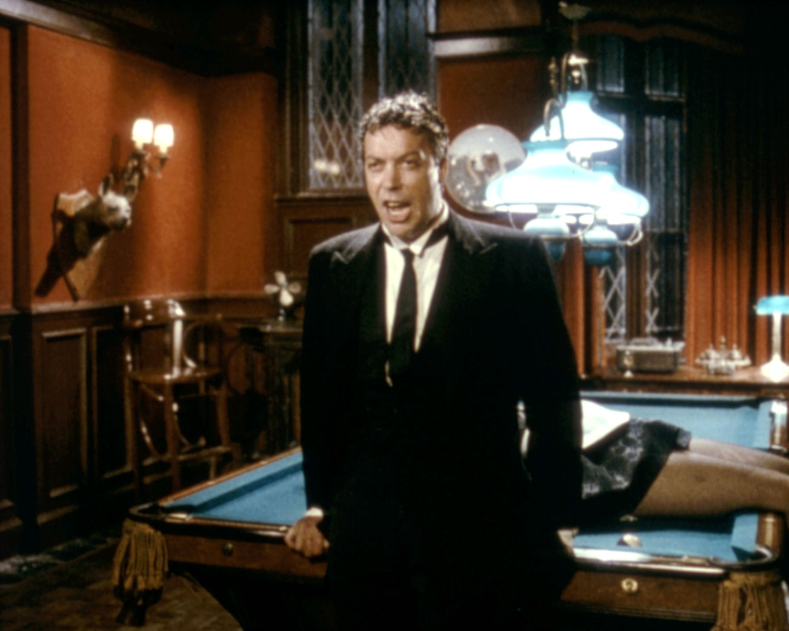 A man in a suit stands next to a pool table in a lavish room, with someone lying on the table behind him