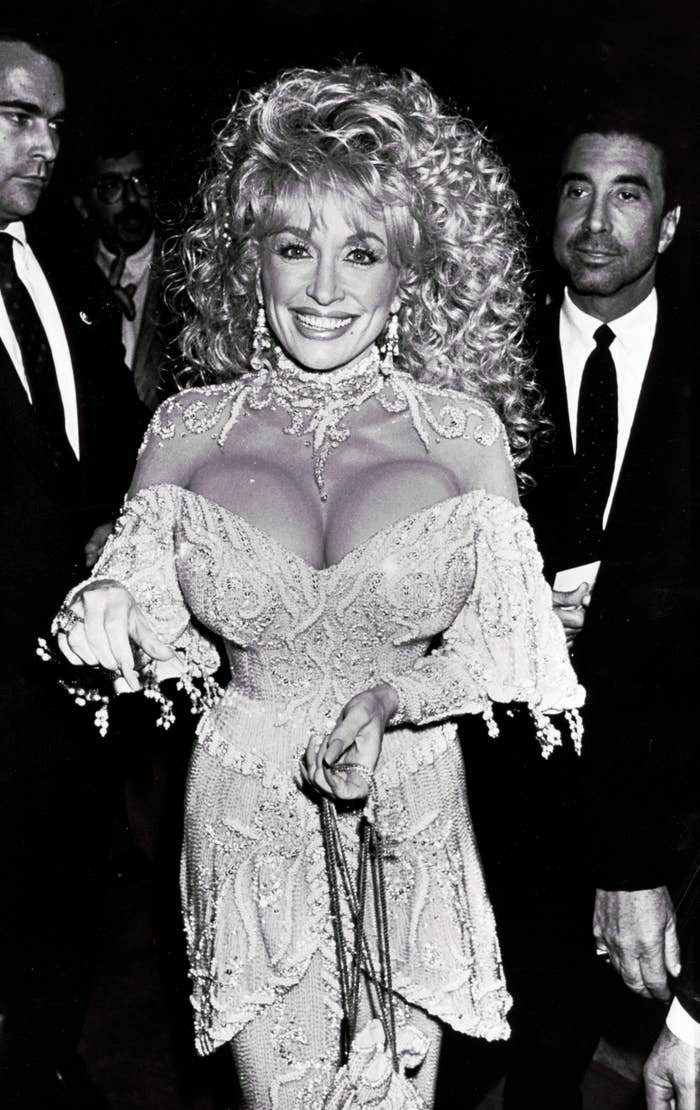 A woman in an ornate, embellished gown with intricate lace details poses on the red carpet, smiling at the camera
