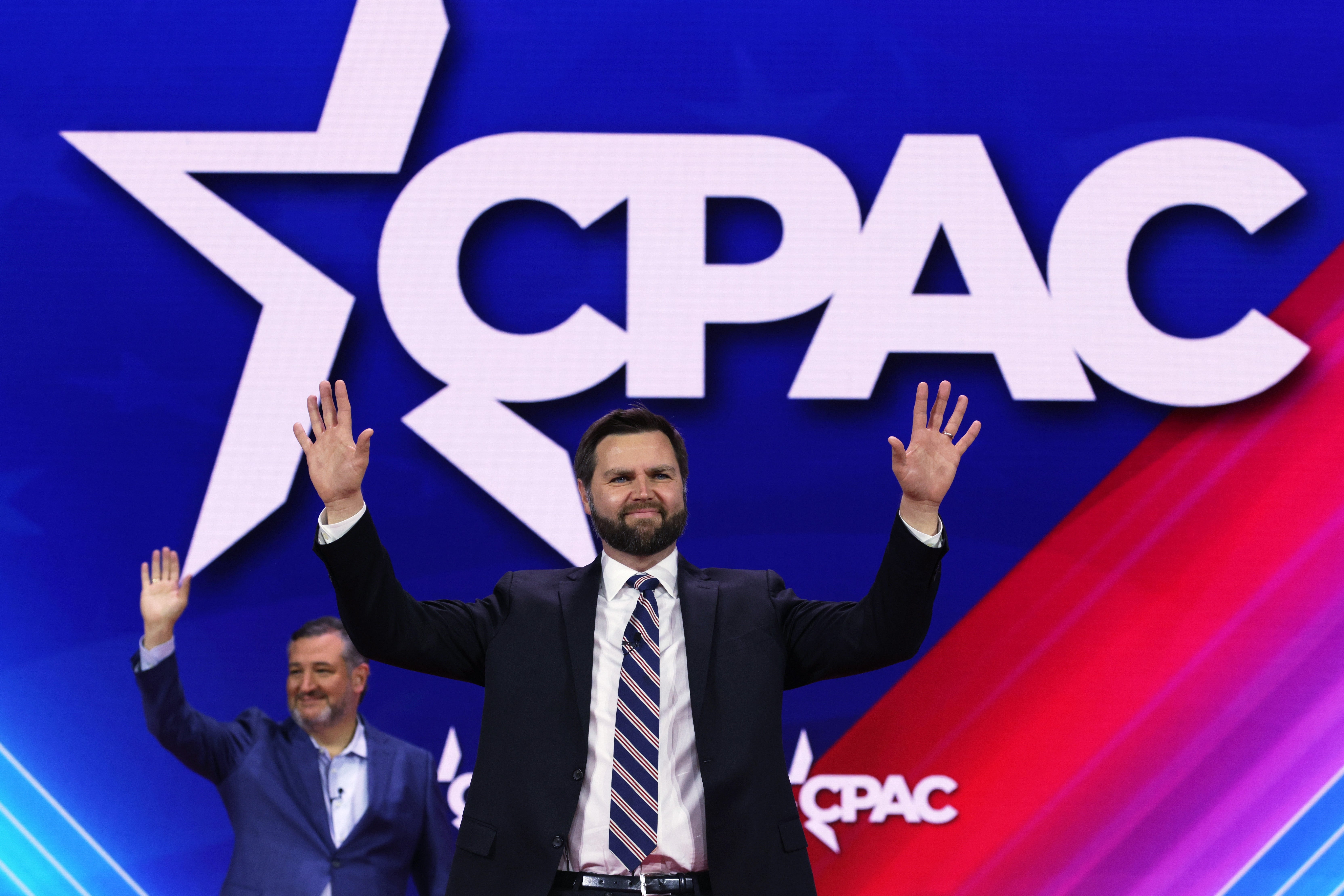 To menn i dress vinker på scenen på CPAC, en konservativ politisk begivenhet, foran en stor CPAC-logobakgrunn