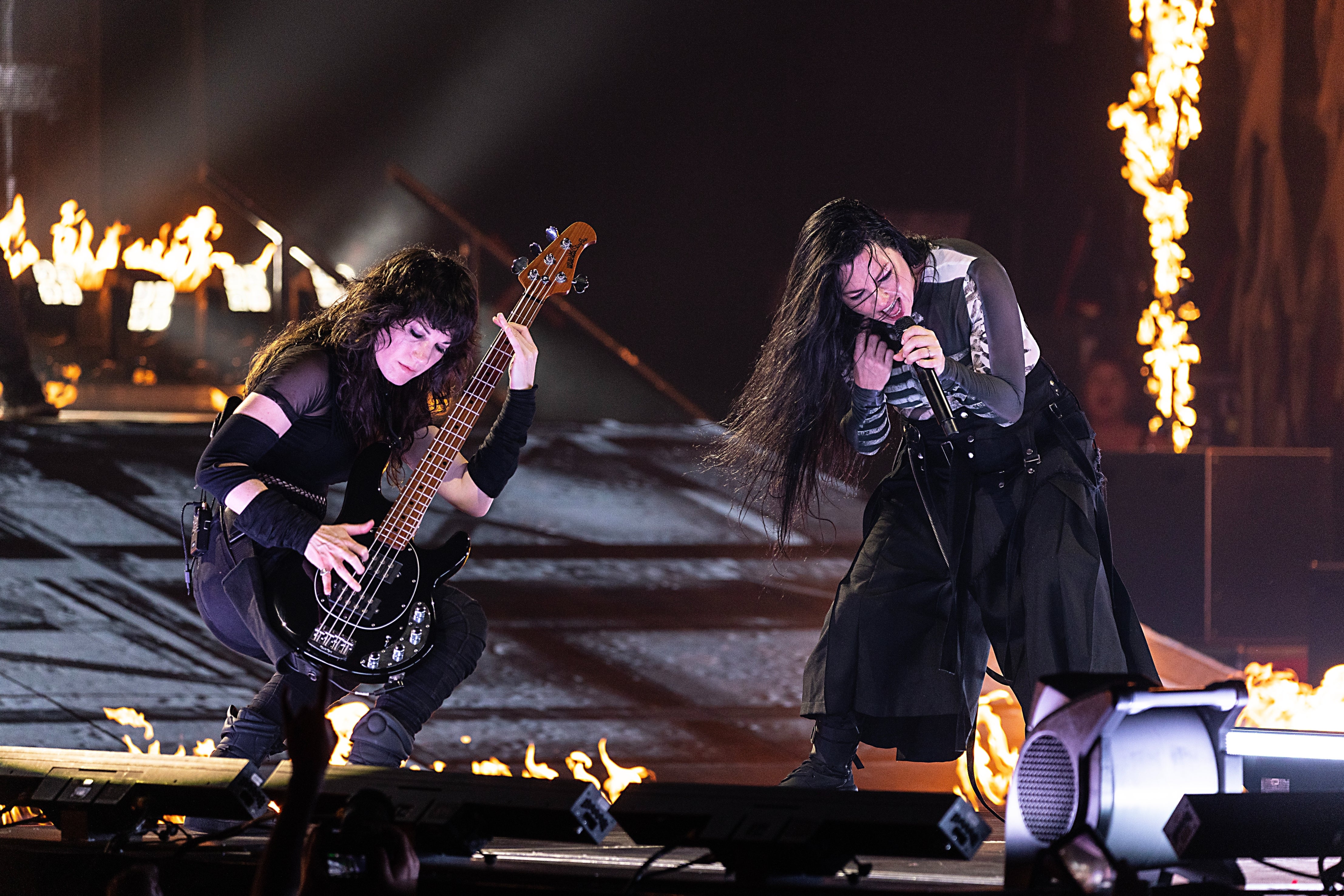 Two performers energetically playing guitar and singing on stage, surrounded by dramatic flames, during a live music concert