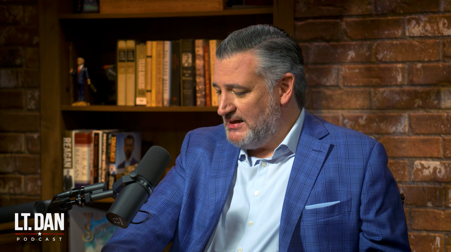 Mann i blå blazer snakker inn i en podcastmikrofon, sittende i et rom med bokhyller