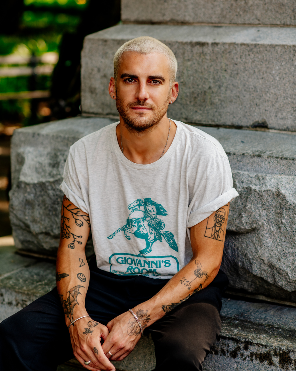 Person with short hair, wearing a t-shirt with "Giovanni's Room" text, sitting on stone steps. Visible tattoos on arms