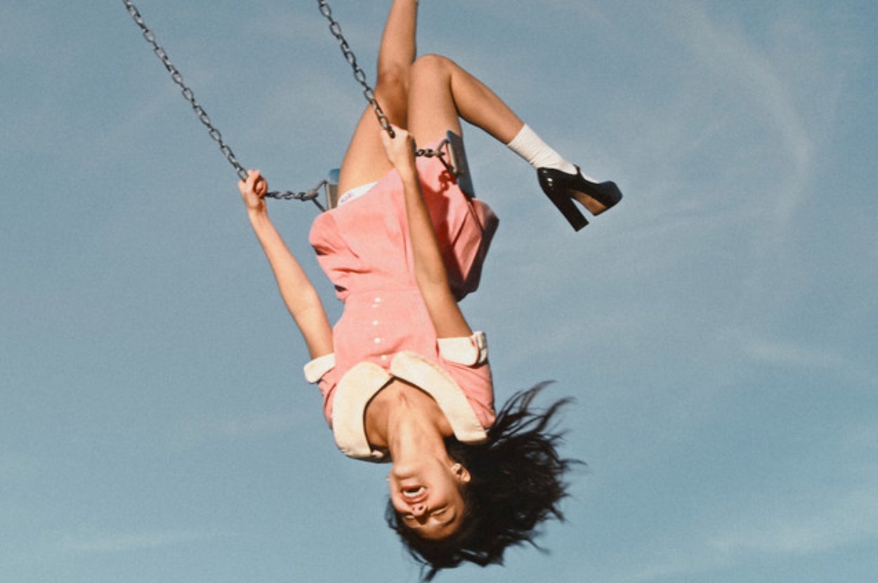 Olivia Rodrigo joyfully swinging upside down in a dress with a collar and high heels, against a clear sky background on the You Seem Pretty Sad for a Girl So in Love music video