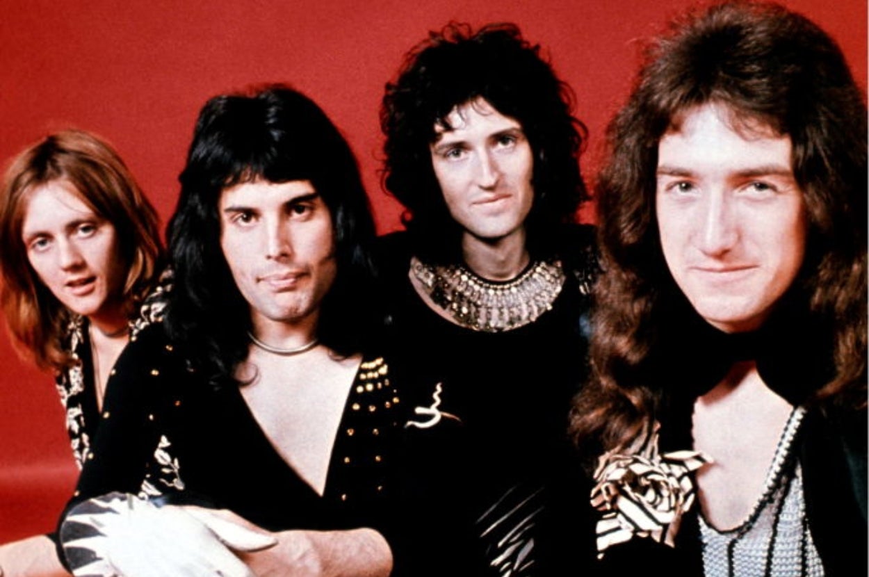 Four members of a 1970s rock band Queen pose in stylish, flamboyant outfits against a plain backdrop