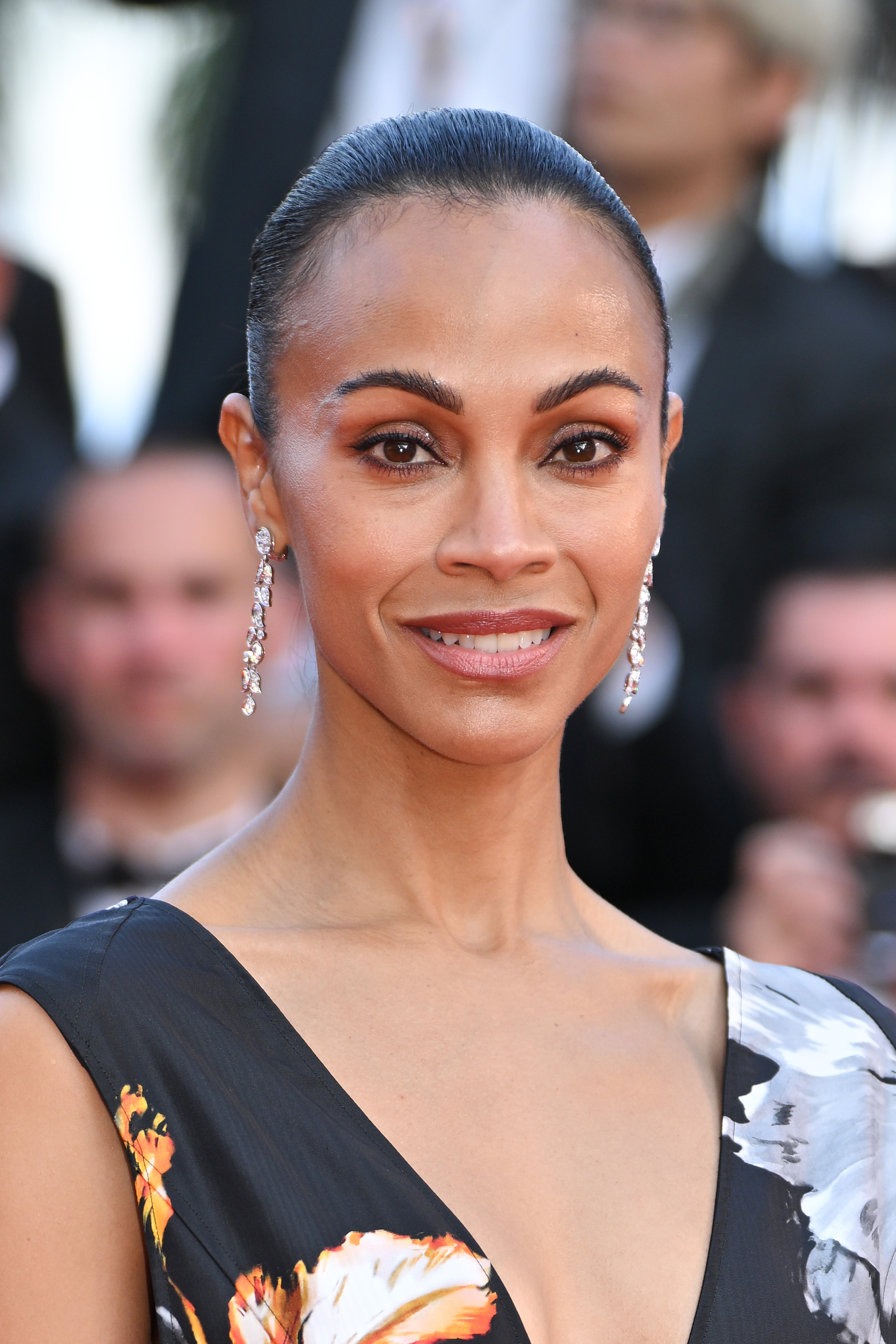 Zoe Saldaña on red carpet in elegant floral dress with deep V-neck, smiling at the camera, and wearing dangling earrings
