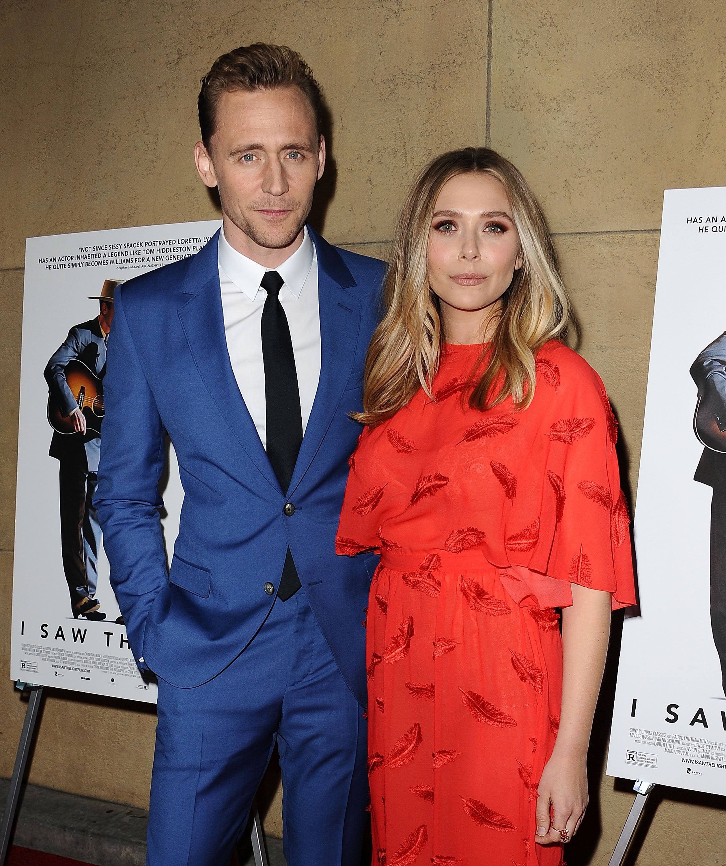 Tom Hiddleston and Elizabeth Olsen pose on the red carpet; Tom in a blue suit, Elizabeth in a red dress, in front of movie posters featuring a guitar player