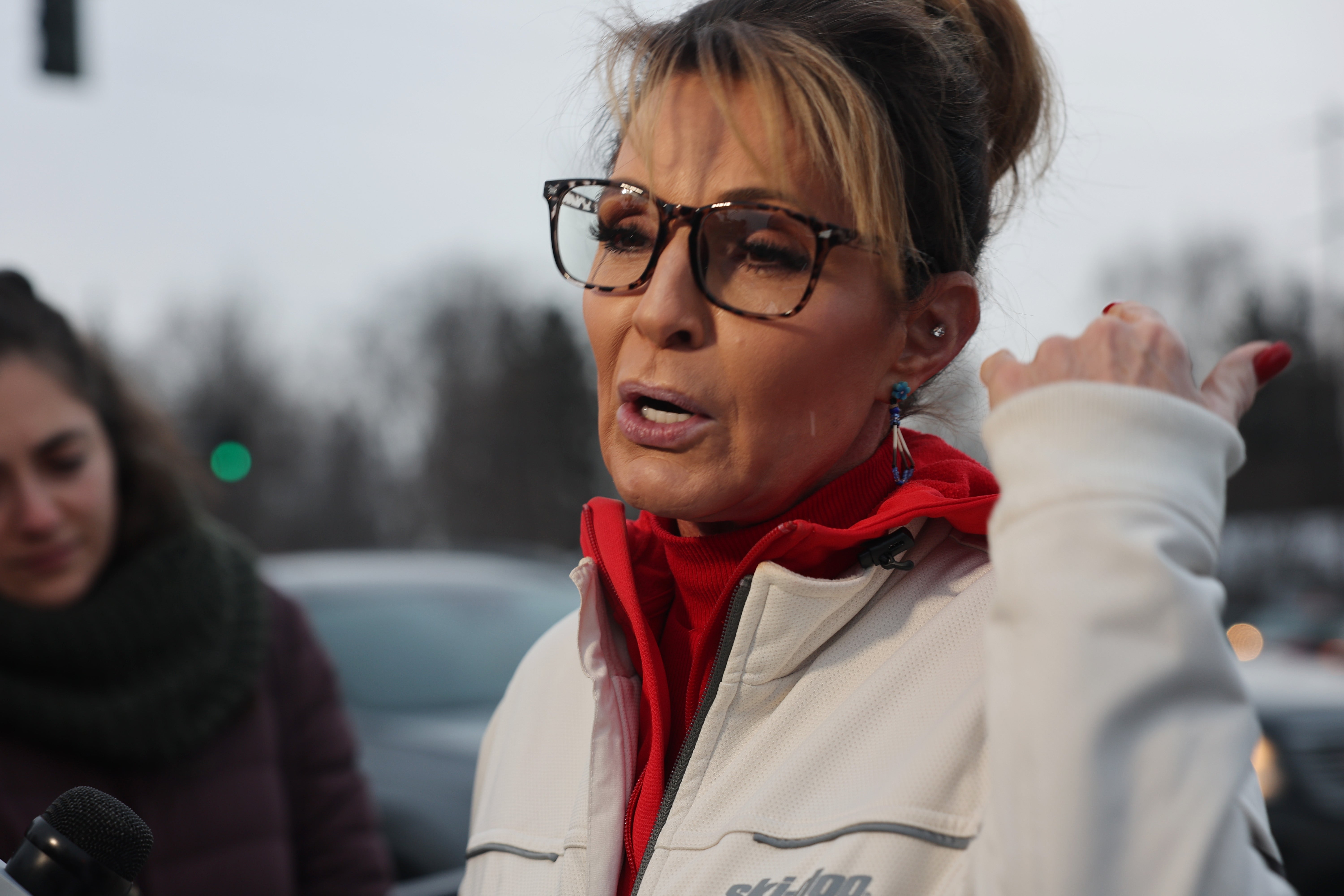 Sarah Palin outdoors speaking to reporters, wearing glasses, a red turtleneck, and a light jacket