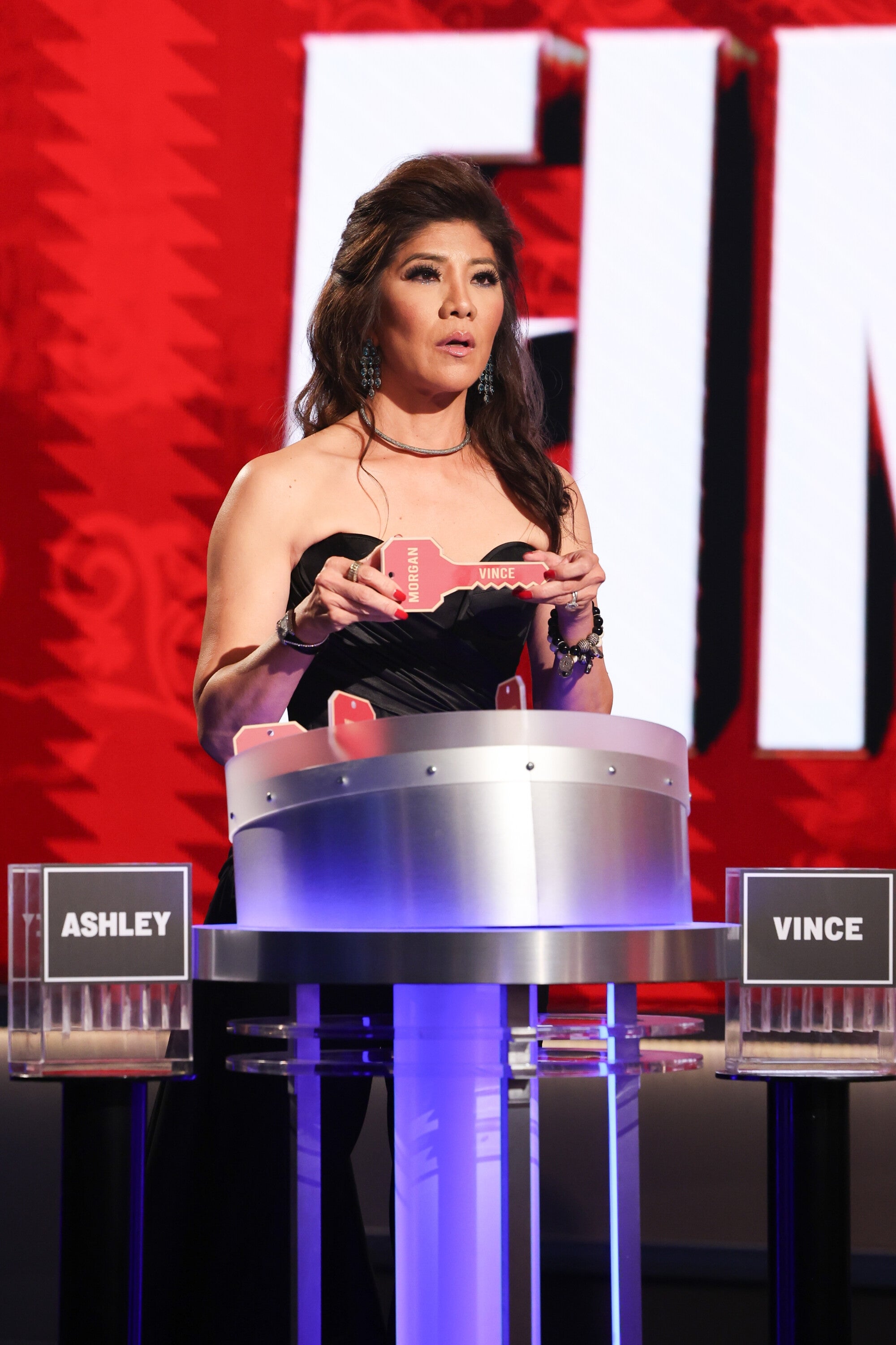 Person in a formal, strapless black dress stands at a game show podium, holding a key marked "Vince," with "FIN" partially visible behind