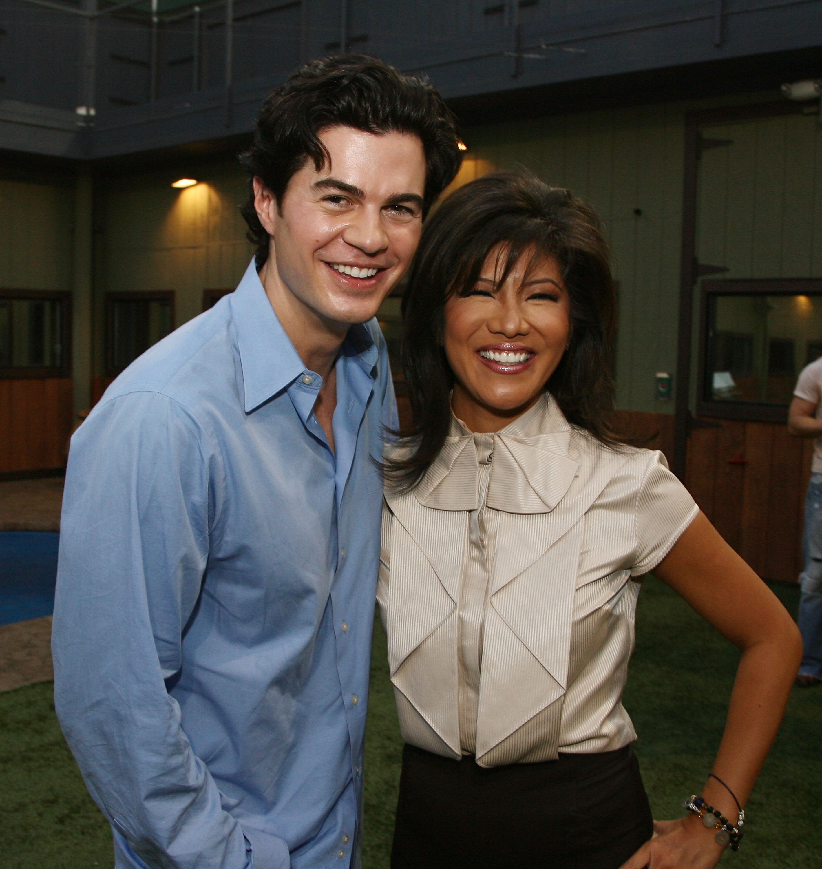 Two people smiling, standing close together. The man is in a casual button-up shirt, and the woman is wearing a stylish blouse with a high collar