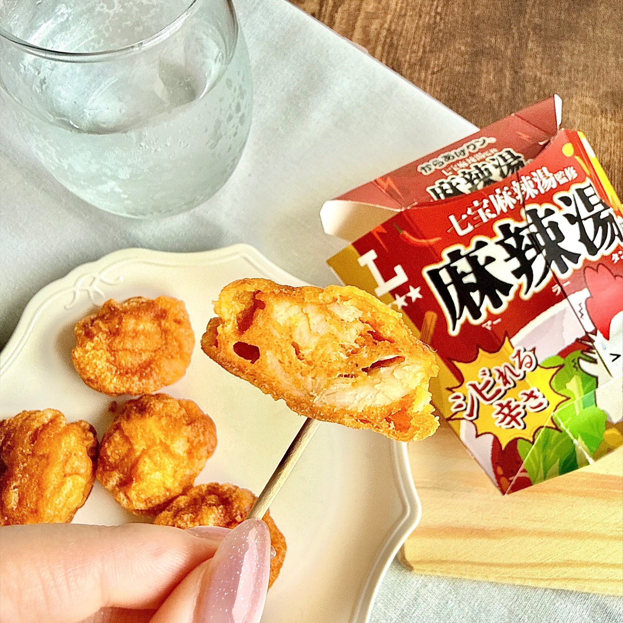 手に持ったローソン 「からあげクン 七宝麻辣湯監修 麻辣湯味」の断面と、皿に盛られた複数のからあげクン、パッケージと飲み物がテーブルに置かれている。