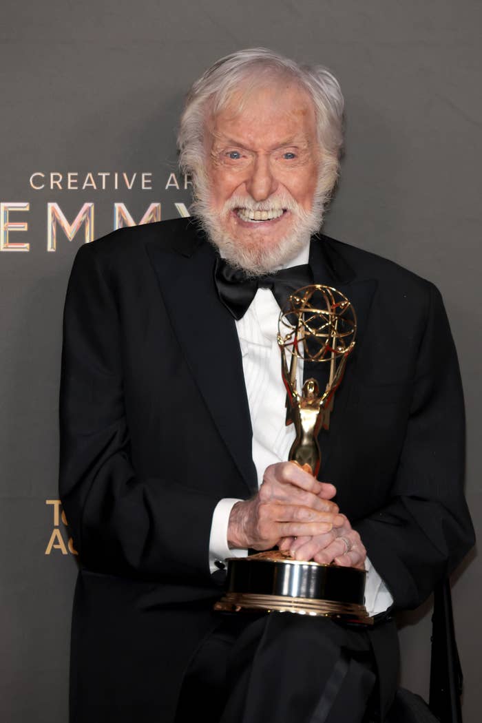 Elderly man in a classic tuxedo holding an Emmy award, smiling broadly