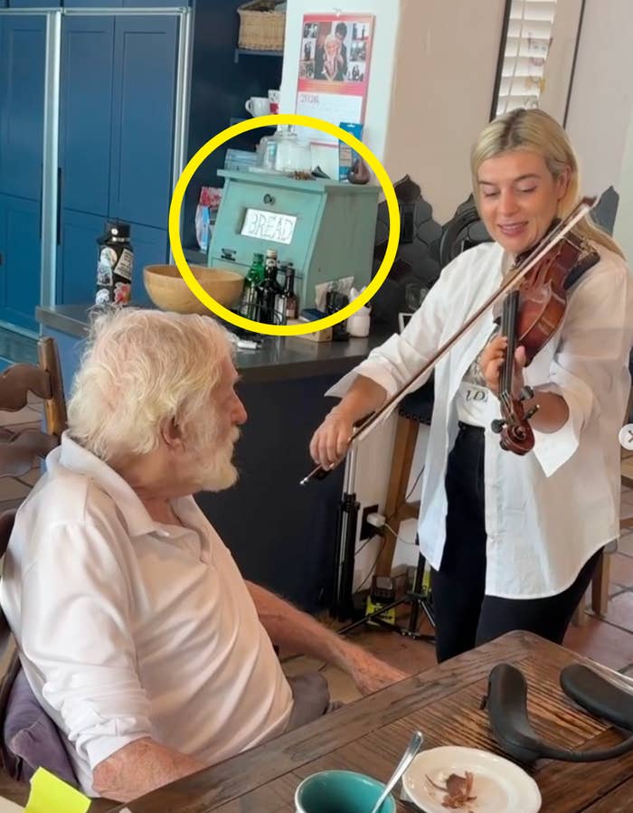 An elderly man sits at a table while a woman plays the violin for him in a cozy kitchen setting