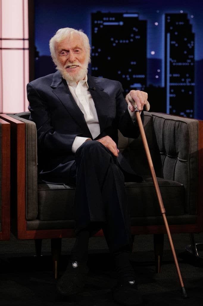 A smiling older man with a beard, in a dark suit, holds a walking stick while seated in a studio setting