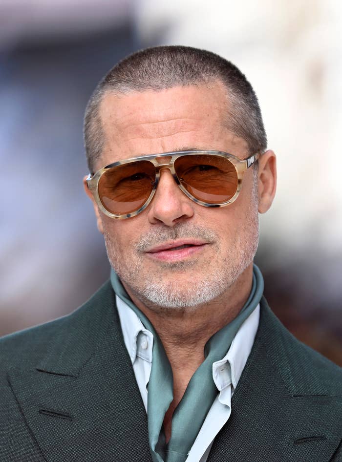 Brad Pitt poses against a blurred background. He wears tortoise-shell aviator sunglasses, a teal tie, and a charcoal grey blazer