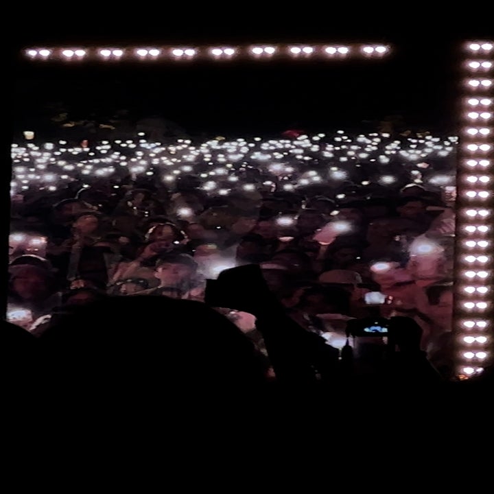 A large crowd holds up lit phones at a concert, creating a sea of lights. Silhouetted figures capture the moment with cameras