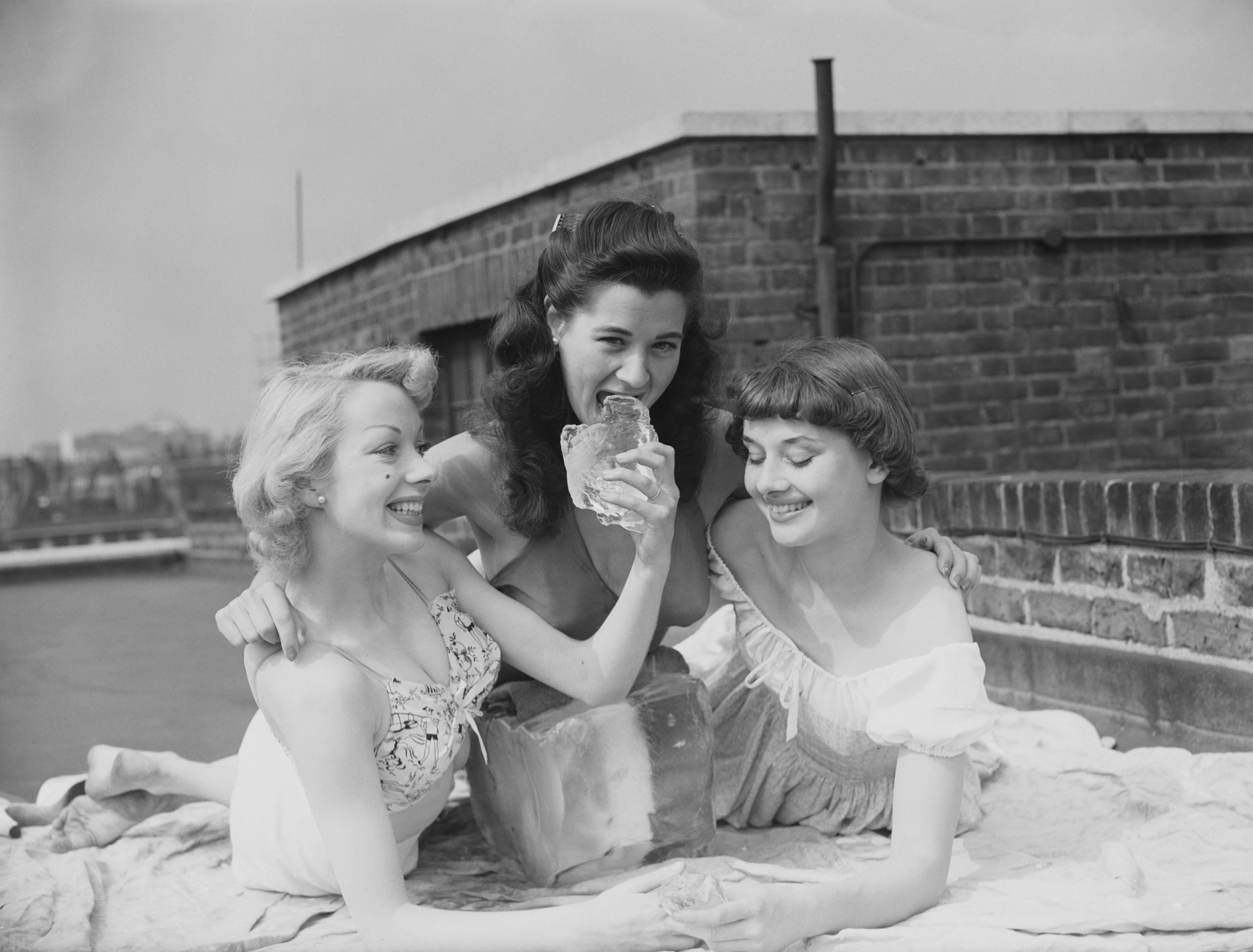 Three women in vintage dresses joyfully pose with a large block of ice on a rooftop, capturing a playful, candid moment