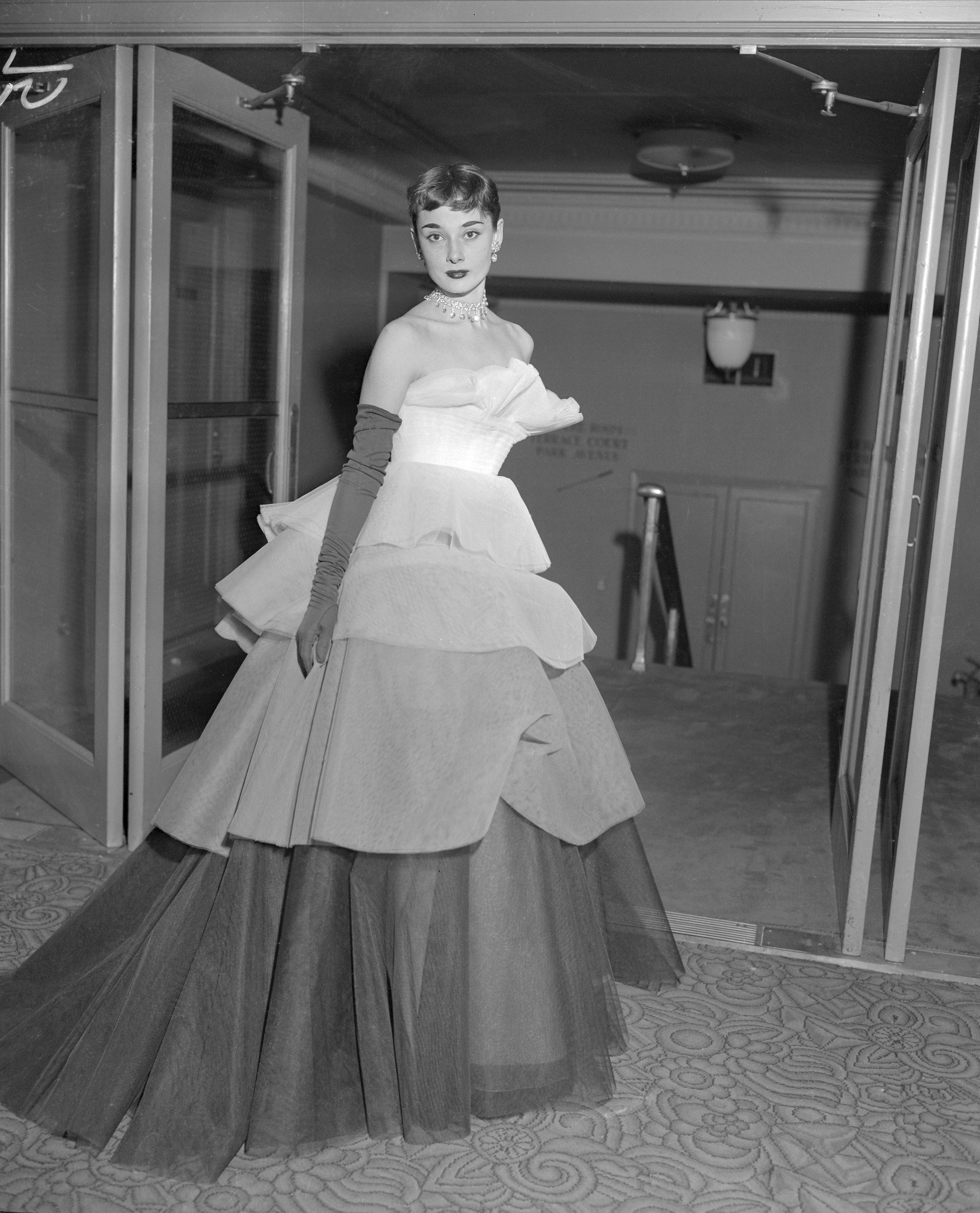 Audrey Hepburn in an elegant, layered gown with gloves, standing gracefully in a hallway